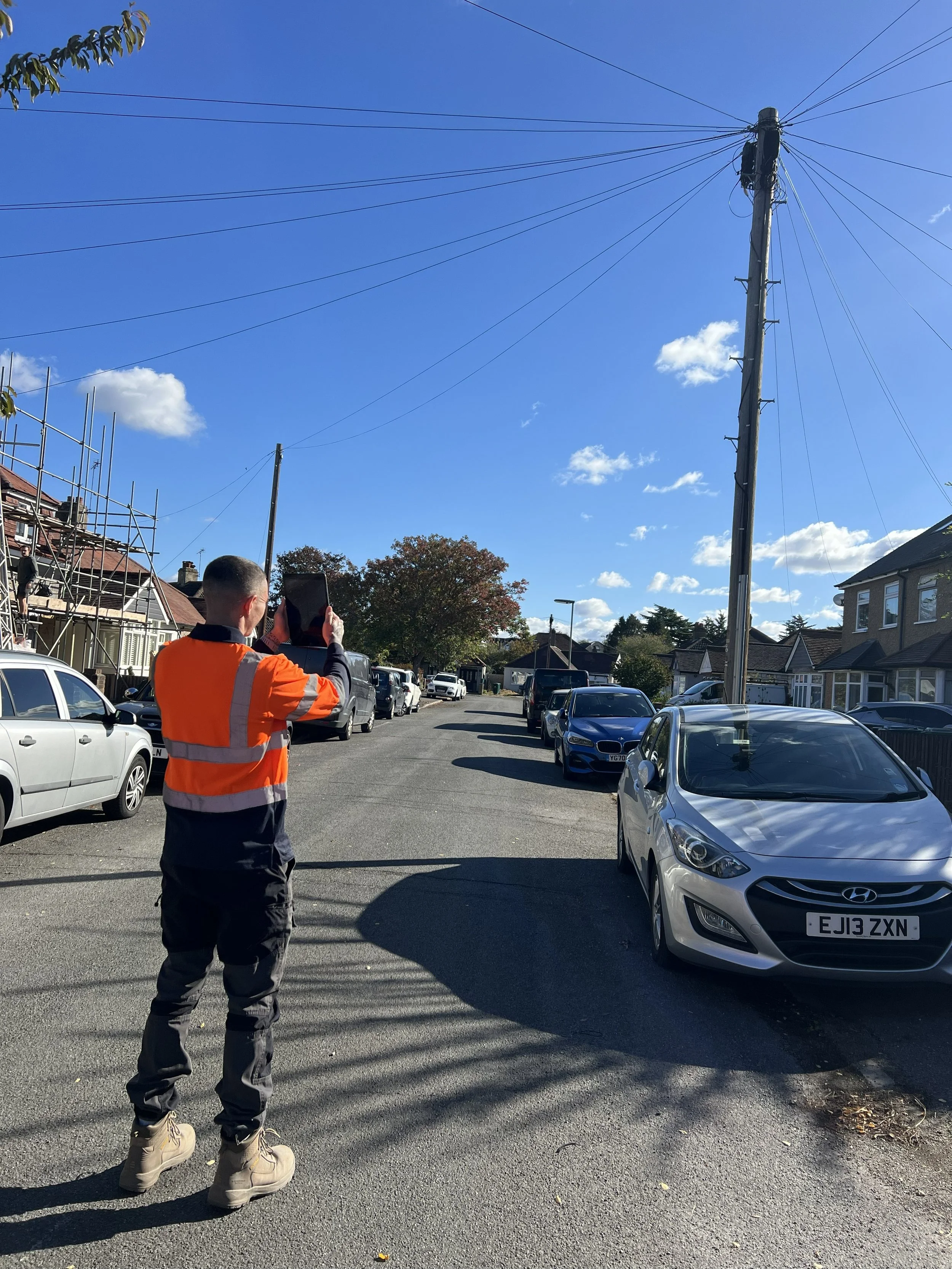 A person wearing an orange safety vest and beige boots is standing on a street, holding a mobile phone and taking a photo or video. The street is lined with parked cars and houses. A utility pole with multiple wires is visible against a blue sky with some scattered clouds.