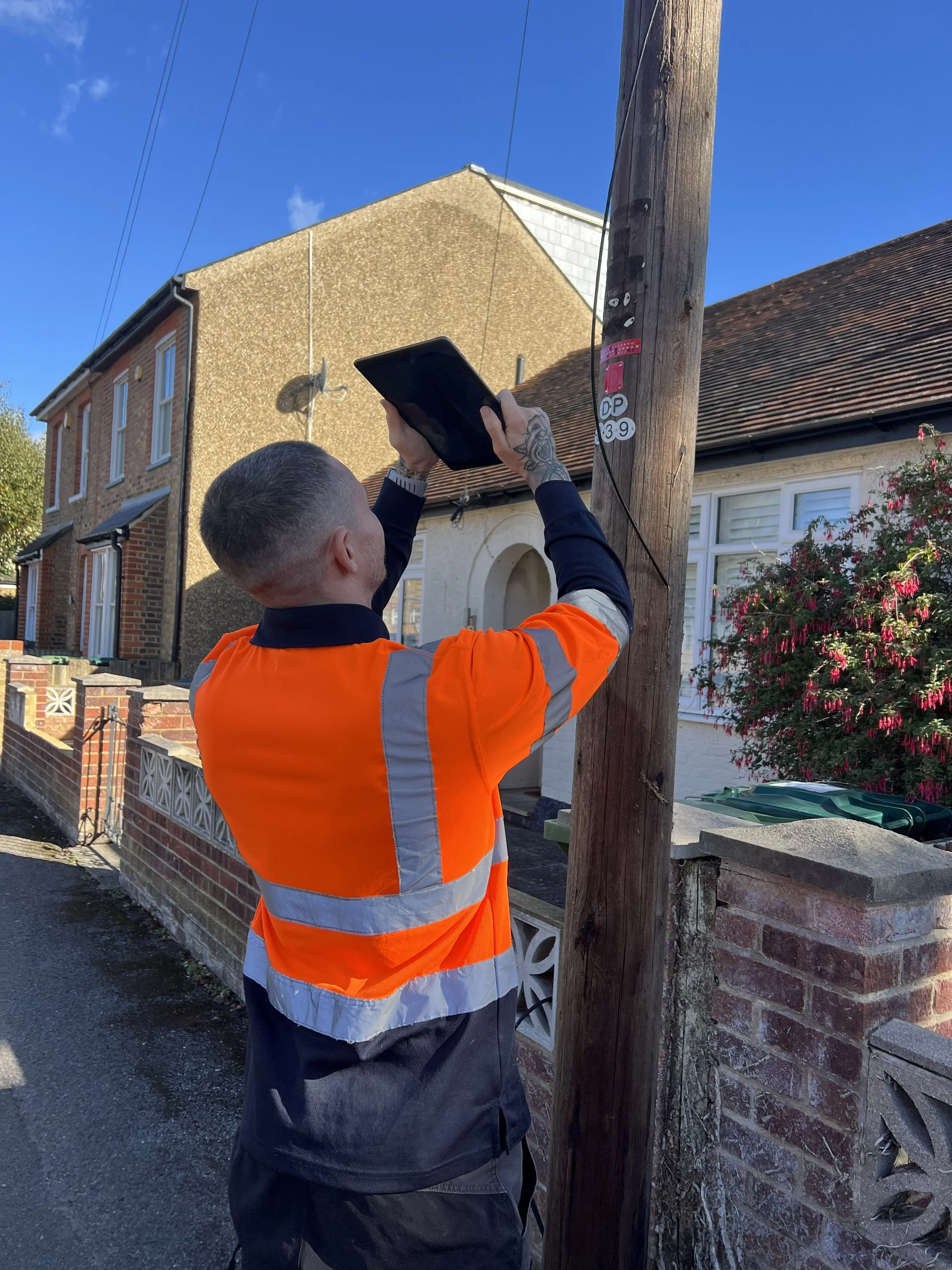 A person in an orange and gray jacket standing on the sidewalk, holding a tablet and working on a utility pole on a residential street.
