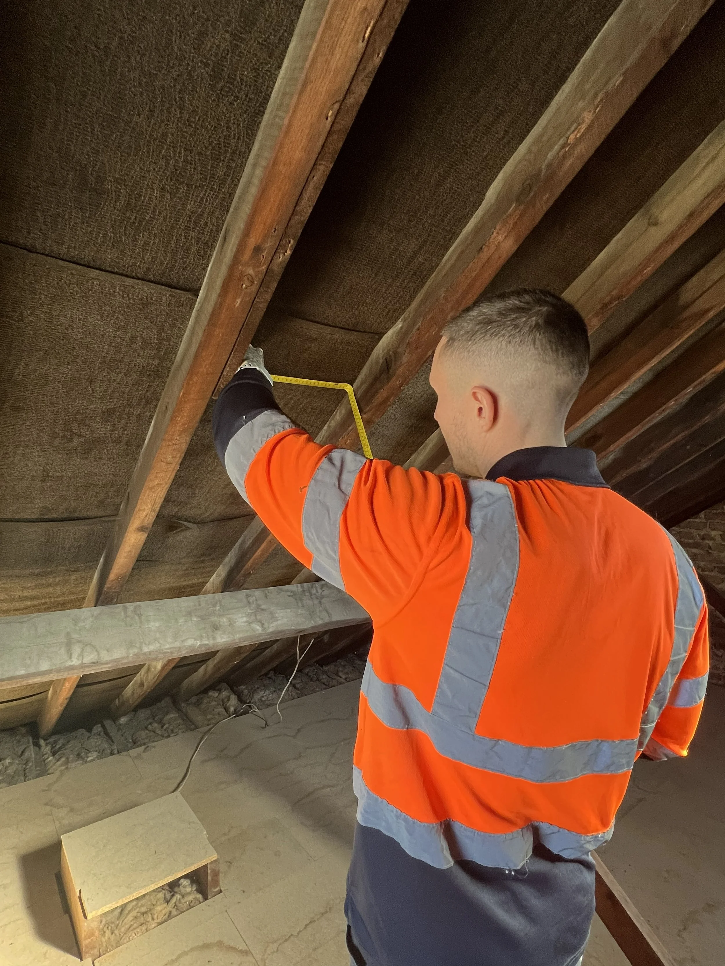 A worker wearing an orange and gray safety jacket using a measuring tape to measure the distance between wooden beams on a ceiling construction site.