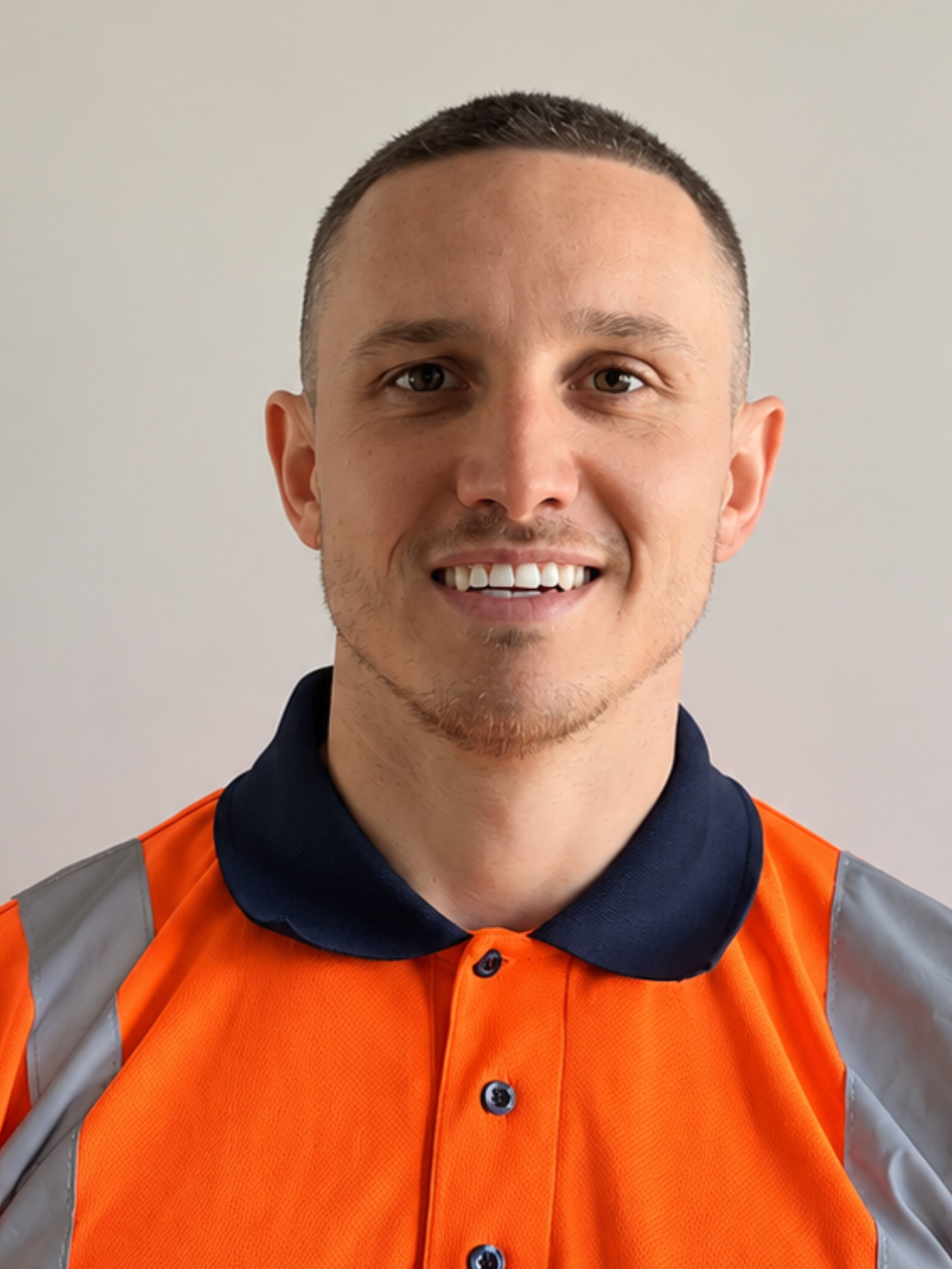 A young man with short dark hair and light skin, smiling, wearing an orange safety vest with gray reflective stripes and a dark polo shirt with a collar.