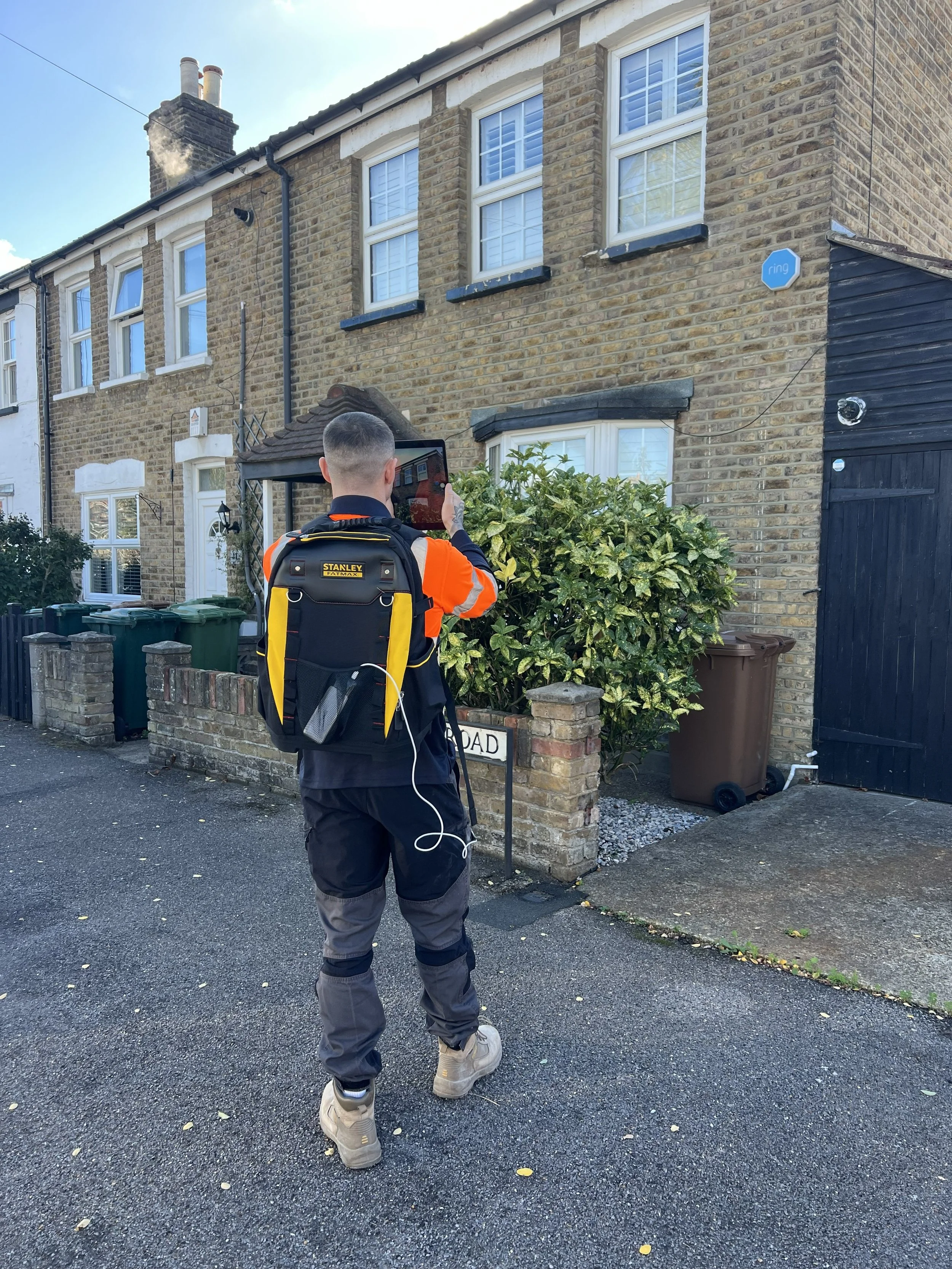 A man wearing a black and yellow backpack and camouflage pants is standing on a paved street in front of a brick house, taking a photo with a tablet. The house has multiple windows with white shutters, a chimney with smoke, and a sign that reads 'ROAD'. There are green and brown trash bins and a bush in front of the house.