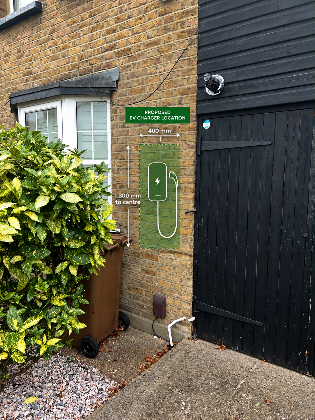 A brick wall with a window and black door, with a marked area for an EV charger installation, including dimensions of 400 mm width and 1300 mm height, and an illustration of an electric vehicle charger. There is a bush covering part of a brown bin, a small pipe on the ground, and outdoor light fixtures.
