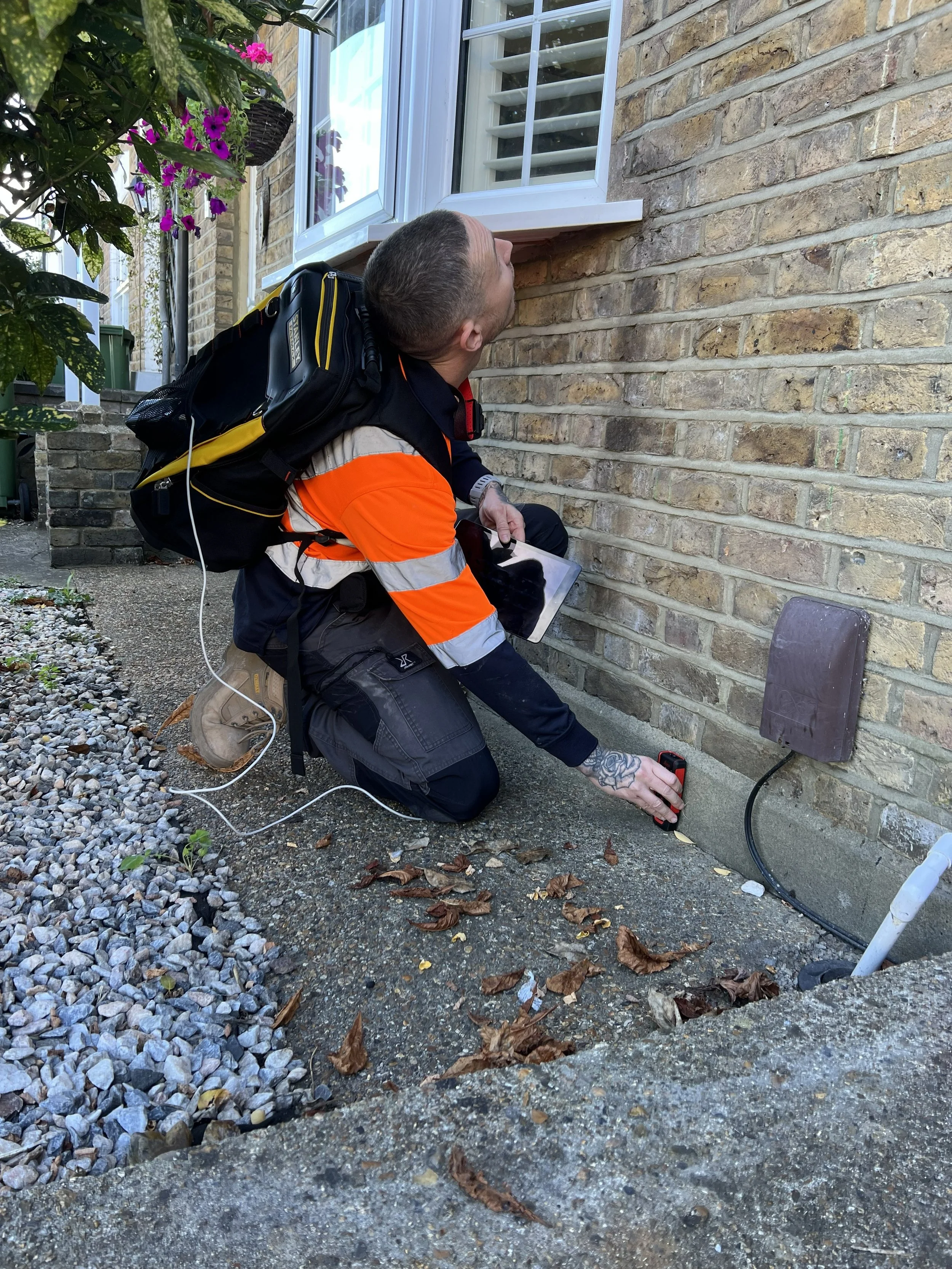 A man in a reflective orange and black uniform kneeling on the ground outside a brick house, inspecting a sewer cleanout with a tool. He has a backpack and a tablet, and is wearing a watch and tattoos on his arm. There are potted plants and purple flowers near the window.