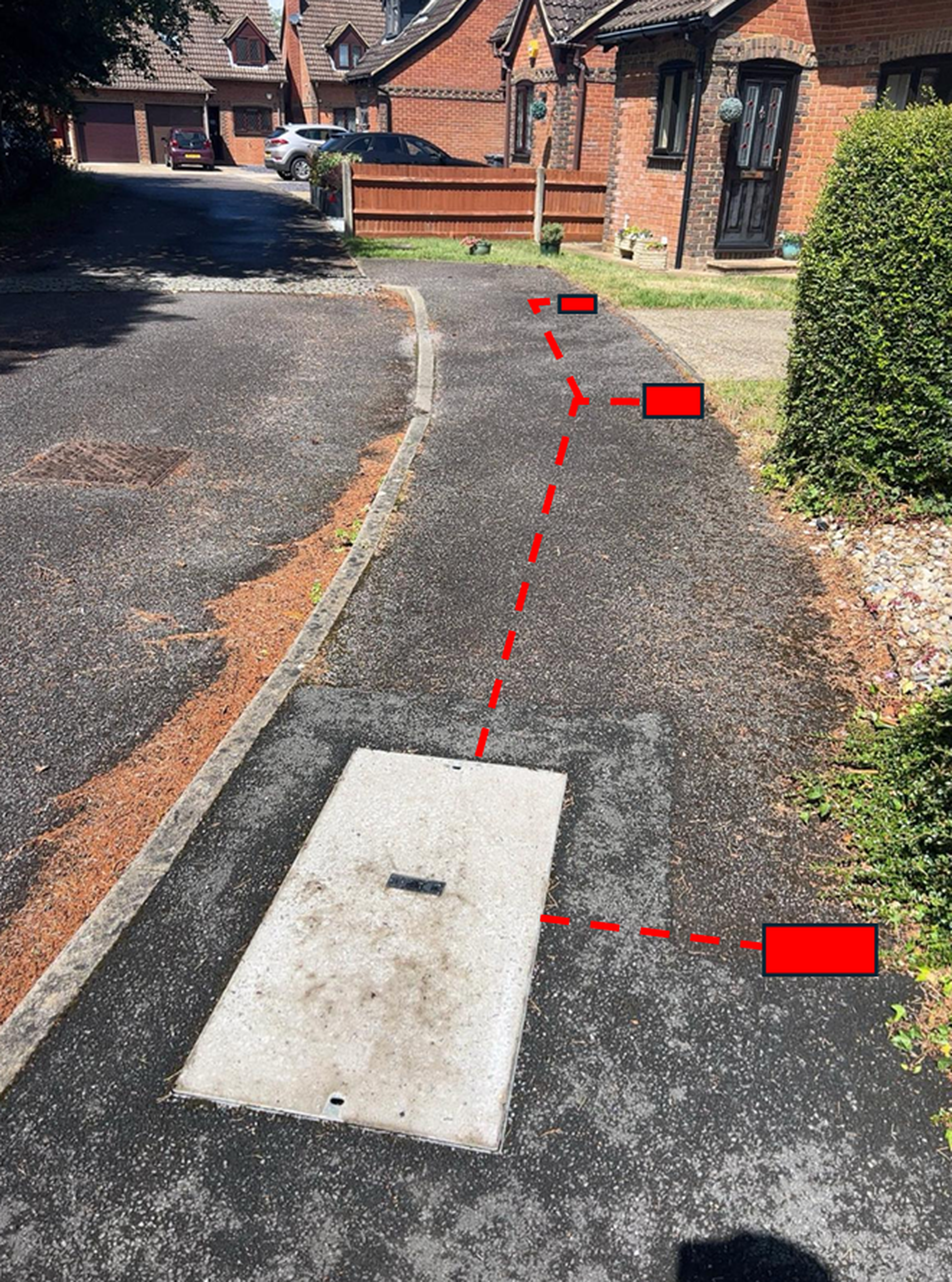 A pathway in a residential area with a missing manhole cover or access panel in the foreground, marked with a red outline and dashed lines, leading toward a street with parked cars and brick houses.