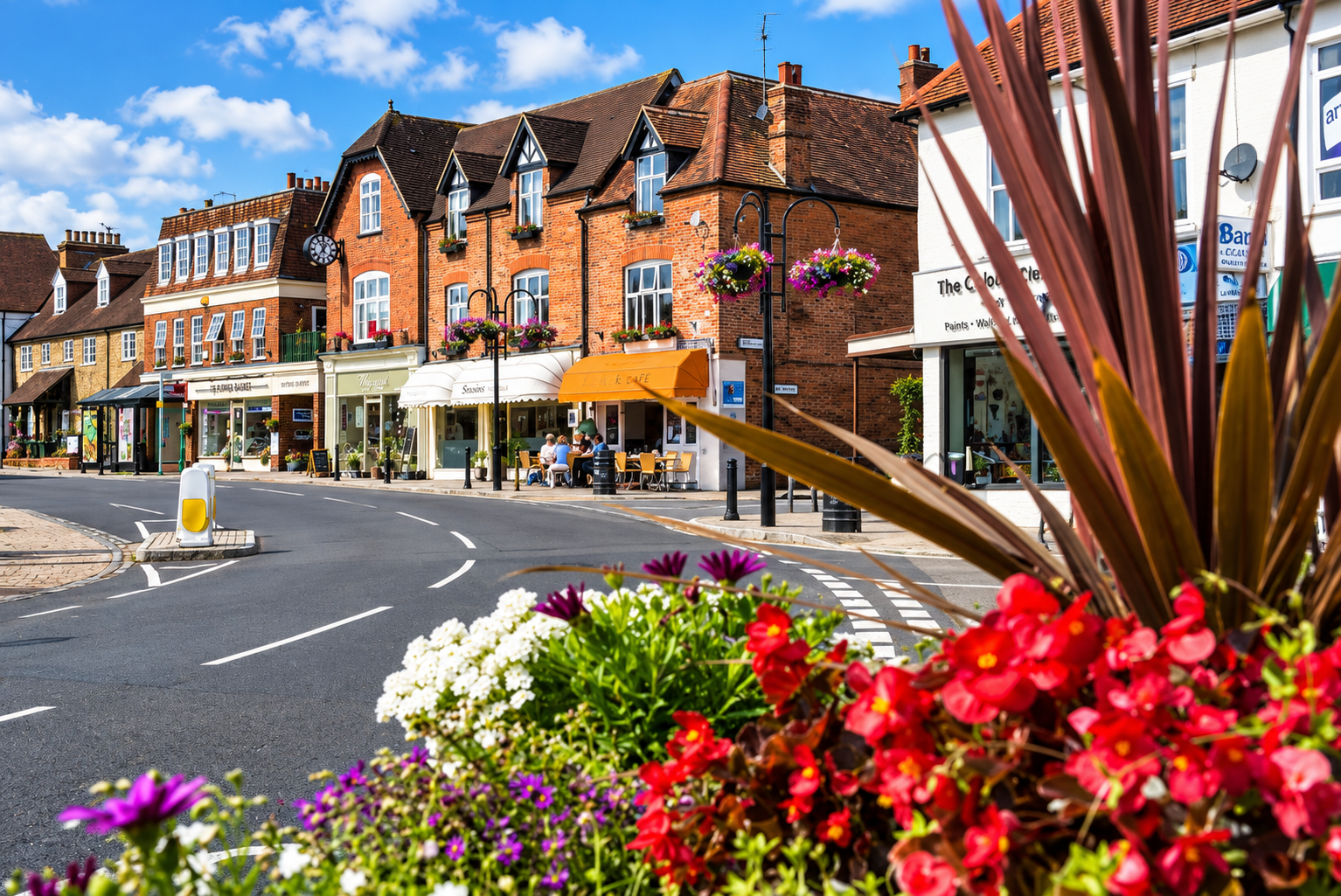 Street view with flower planters, shops, and outdoor seating under a blue sky.