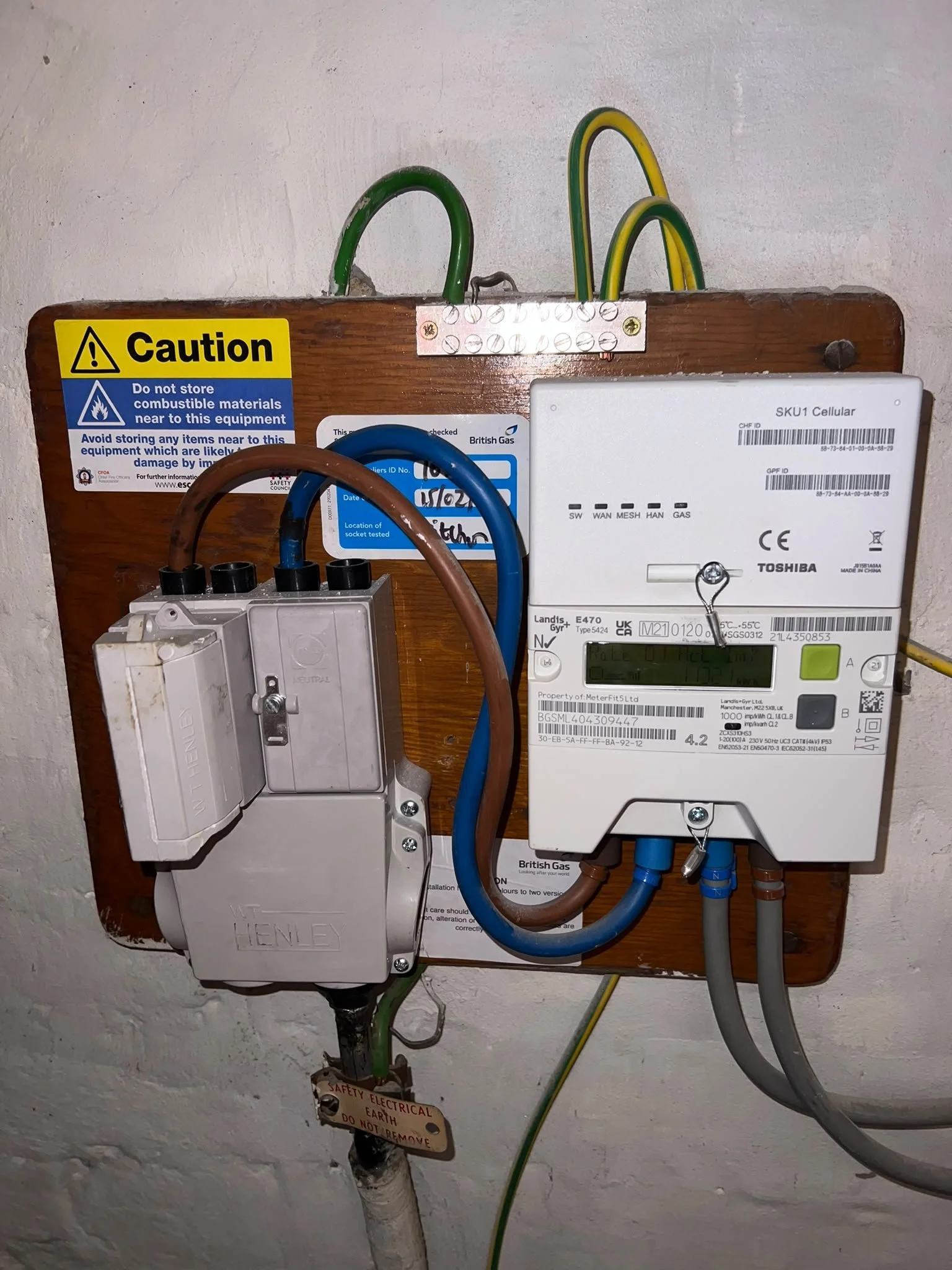 Electrical meters and wiring mounted on a wooden panel attached to a white textured wall, with caution signs and a label indicating electrical safety.