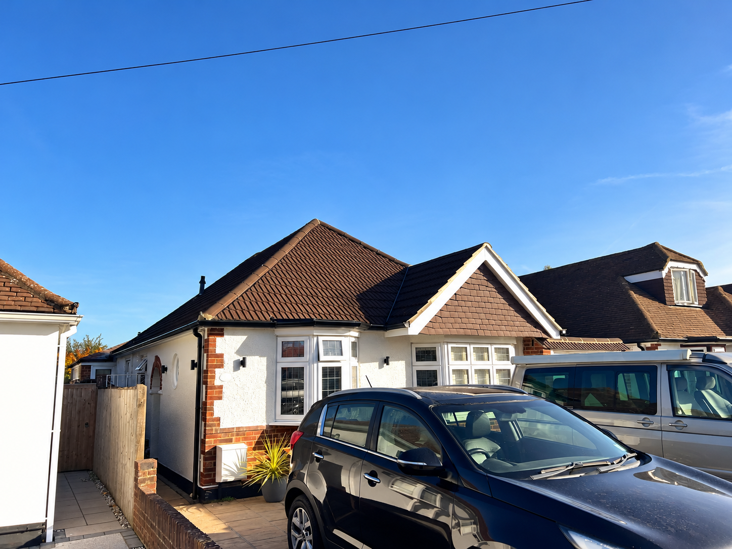 Front view of a white house with a brown tiled roof, black car parked in front, and a van beside it. Clear blue sky.