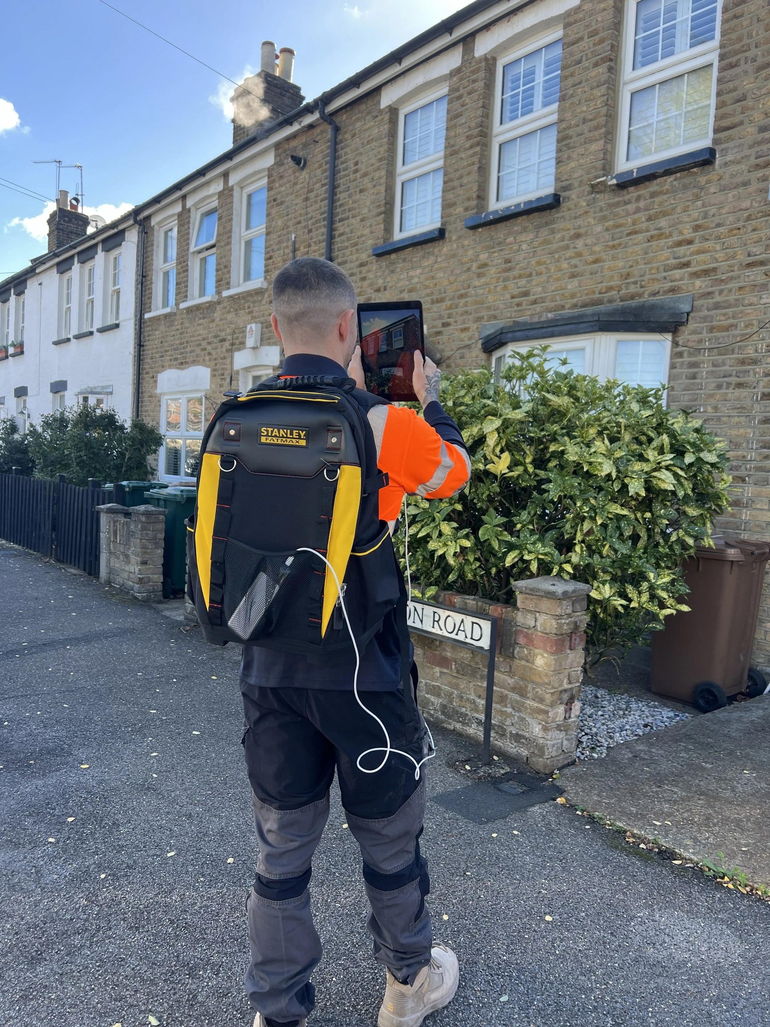 A man wearing a black and yellow Stanley FatMax backpack, orange and black jacket, and grey pants stands on a sidewalk taking a photo of a brick house with his tablet. The house has white-framed windows, a bush in front, and a street sign reading "ON ROAD".