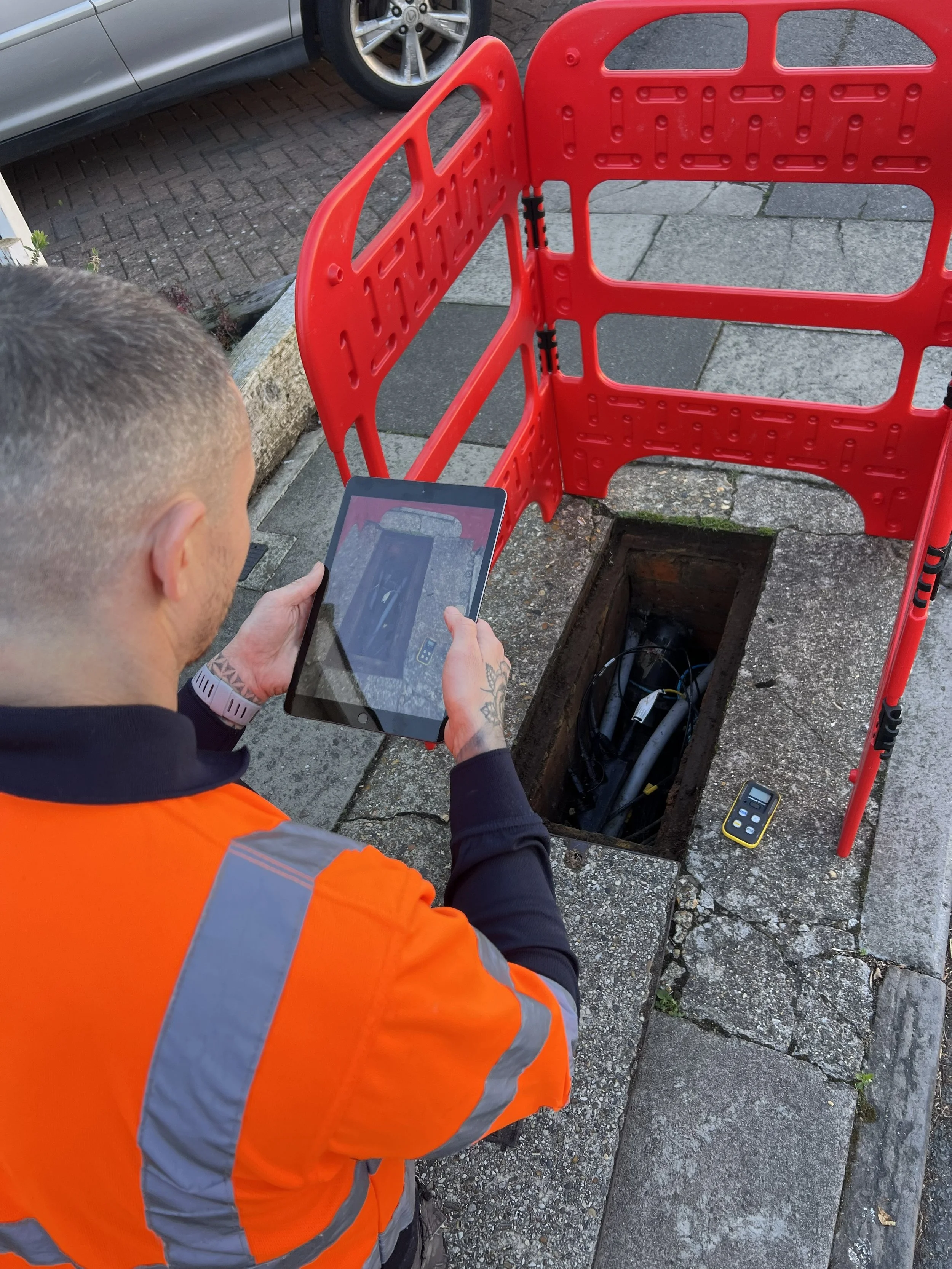 A worker in an orange high-visibility vest and gray long sleeve shirt holding a tablet, inspecting an open manhole on the sidewalk. The open manhole reveals cables and equipment inside, and a remote control device lies nearby on the ground.
