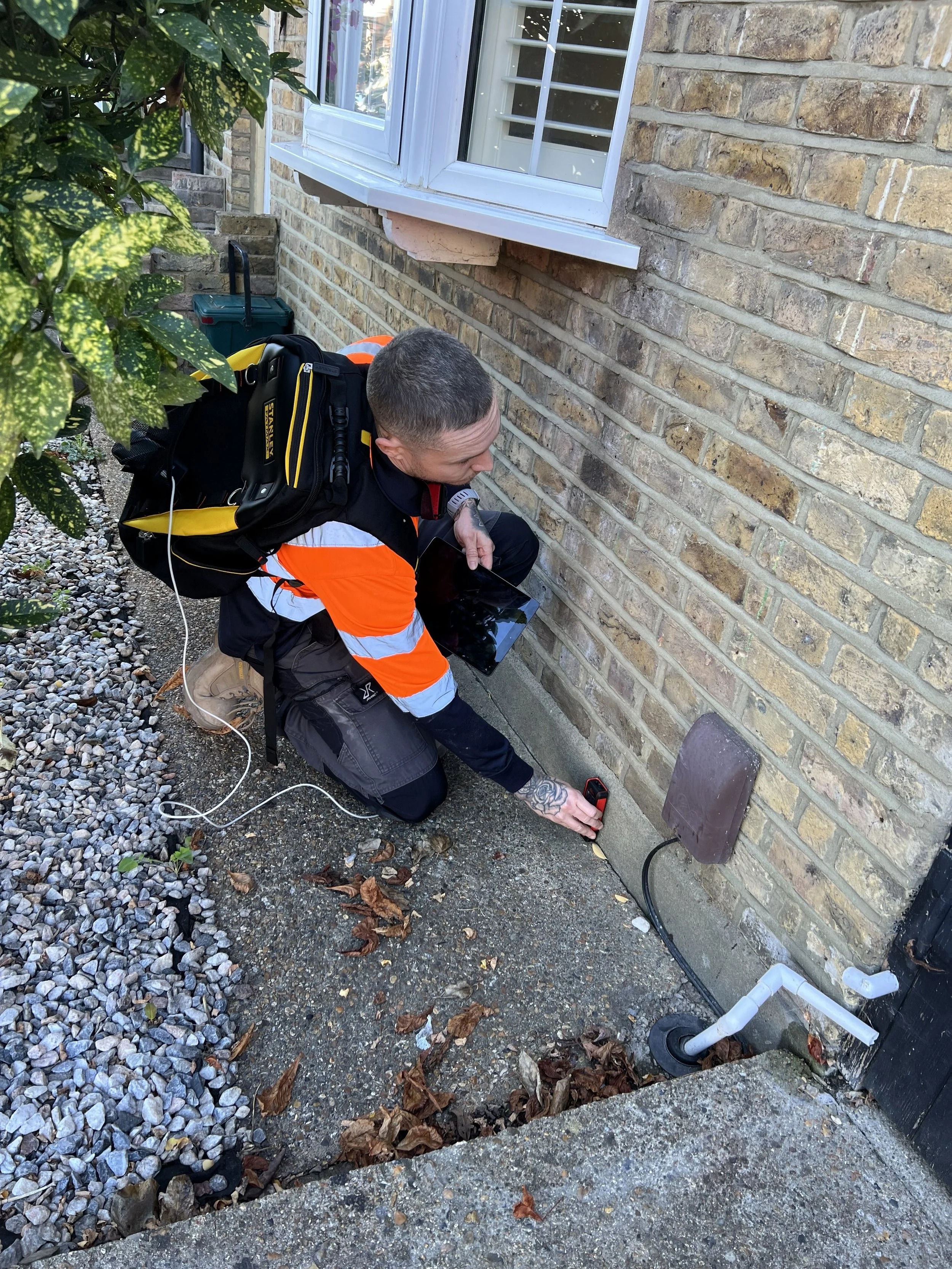 A man with a short haircut and tattoos on his arm kneels outside on gravel next to a brick house, using a laser distance meter to measure the foundation of the house. He is wearing a black and orange safety jacket and carrying a black and yellow backpack.