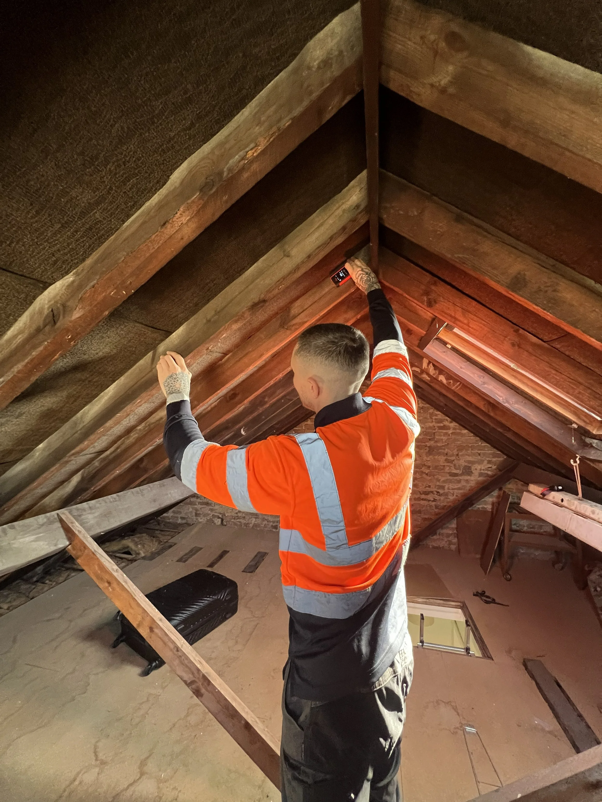 A person wearing an orange safety vest and black clothing is working on an attic ceiling, using a tool to measure or cut the wooden beams.