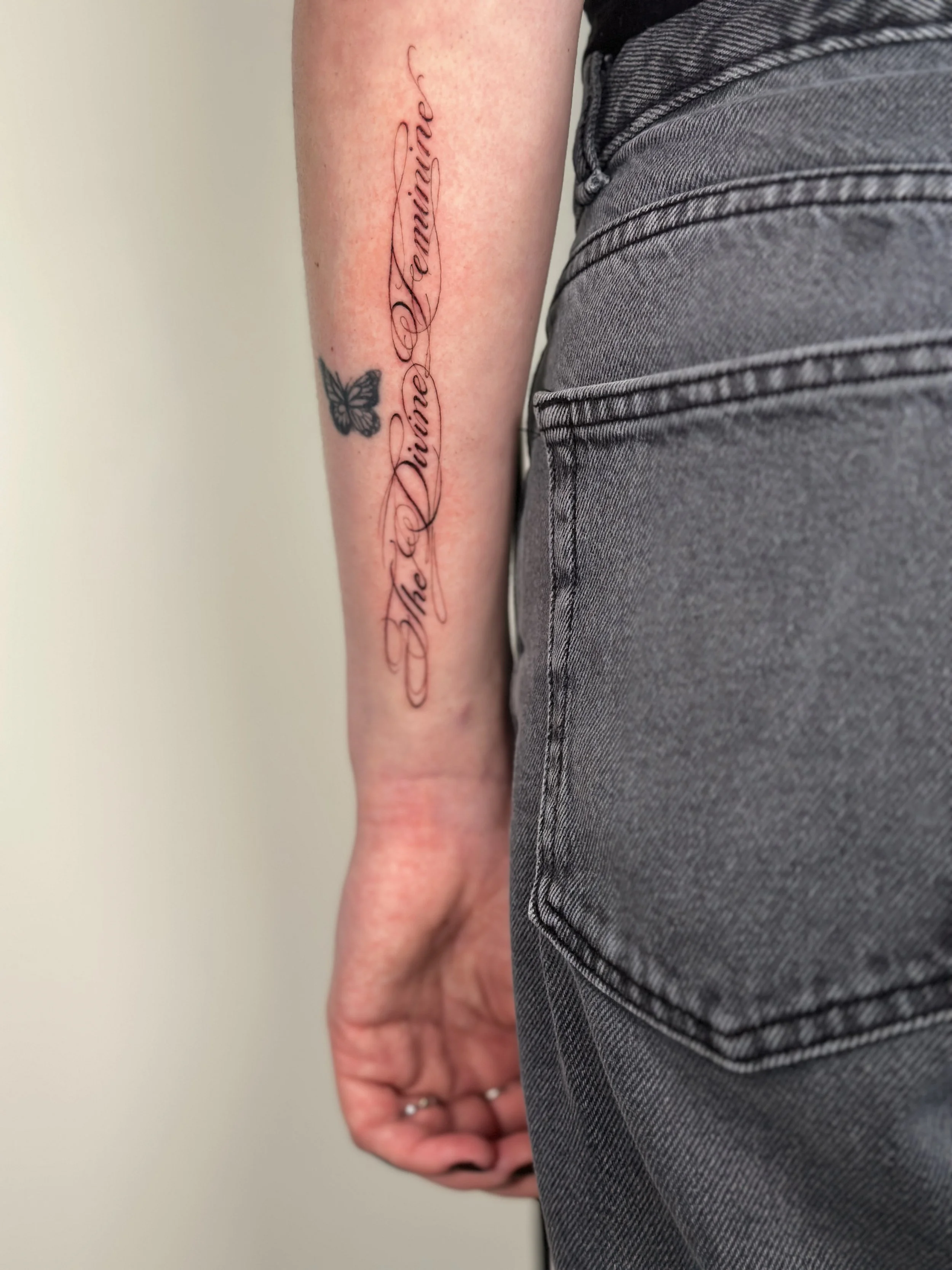 A person's arm with a tattooed phrase 'The Divine Feminine' written in cursive script and a small butterfly tattoo below it, visible beside the person's side in gray jeans.