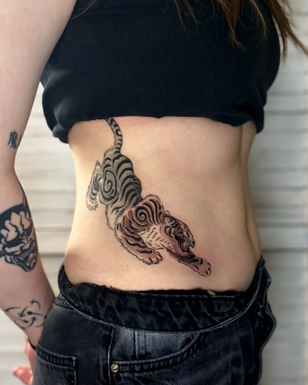 A person with tattoos on their arm and side, wearing a black crop top and black jeans. The side tattoo depicts a stylized korean tiger with swirling black and gray stripes.