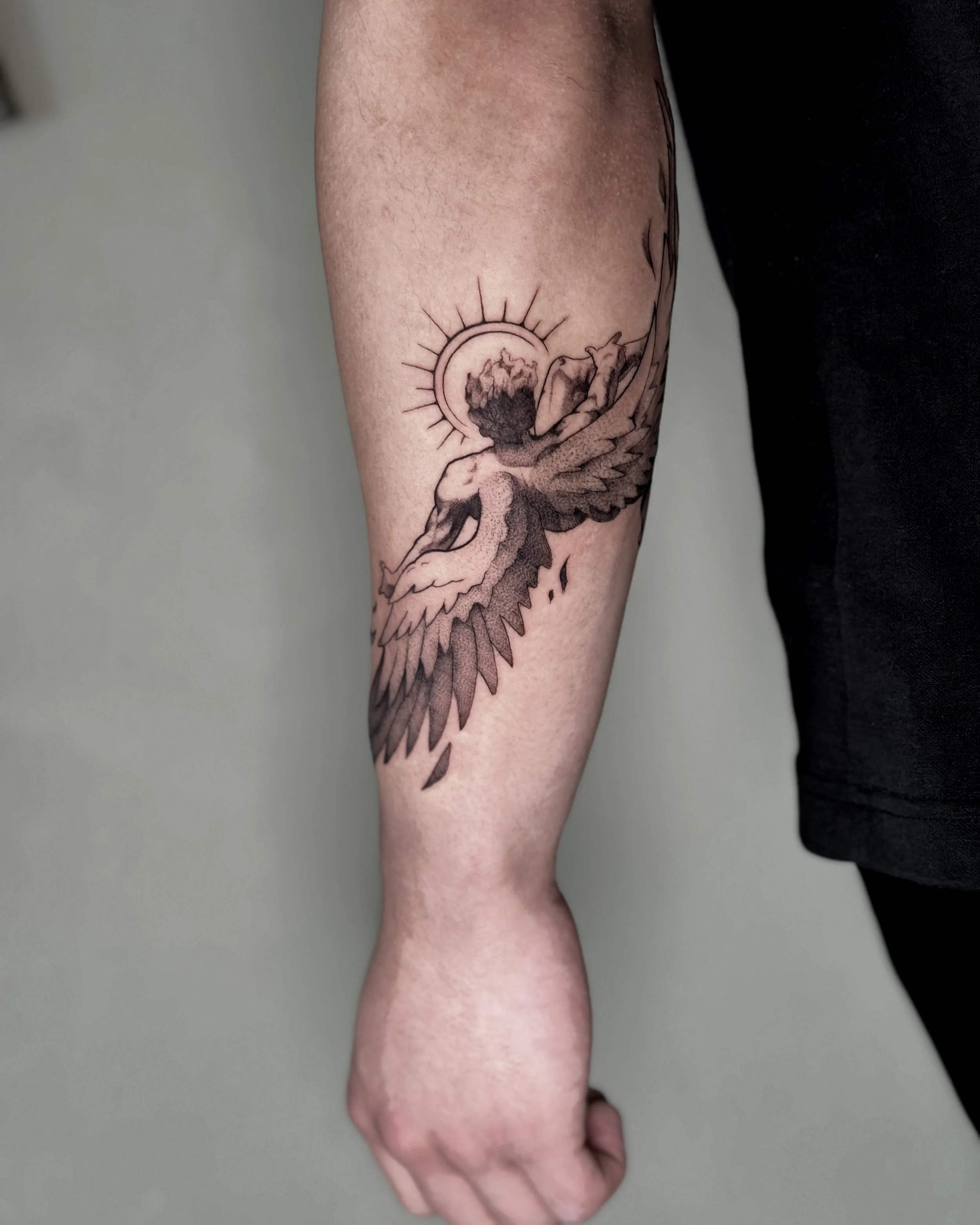 A tattoo of a winged figure with a large circular sun behind it on a person's forearm.