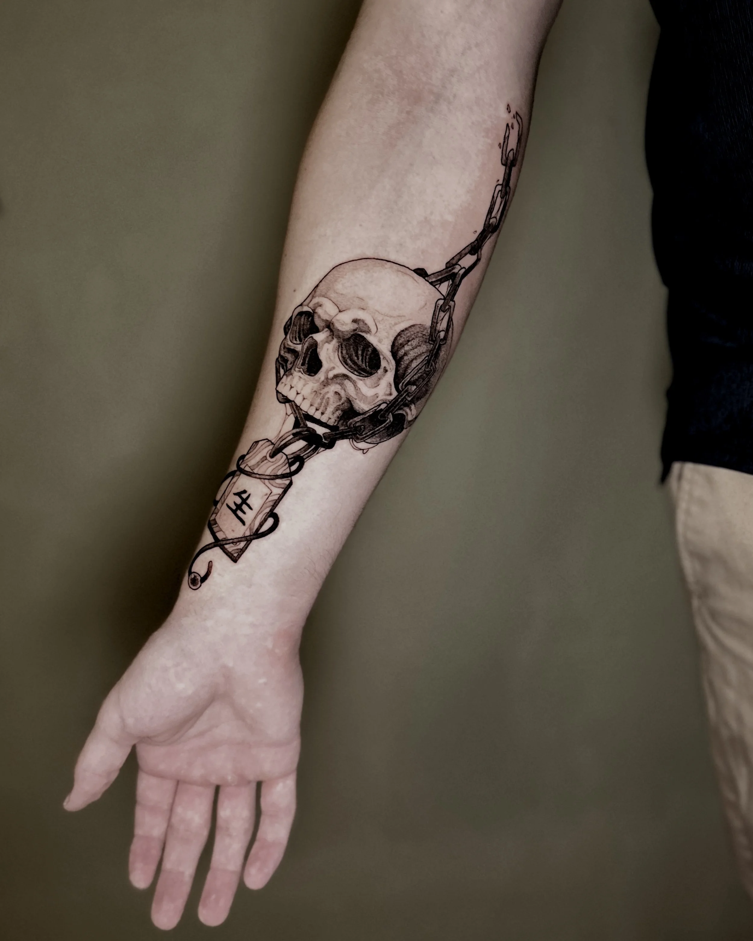 A person's arm with a black line tattoo of a skull with a chain wrapped around it. The person is wearing a black short sleeve shirt, and their hand is extended downward with fingers slightly apart.