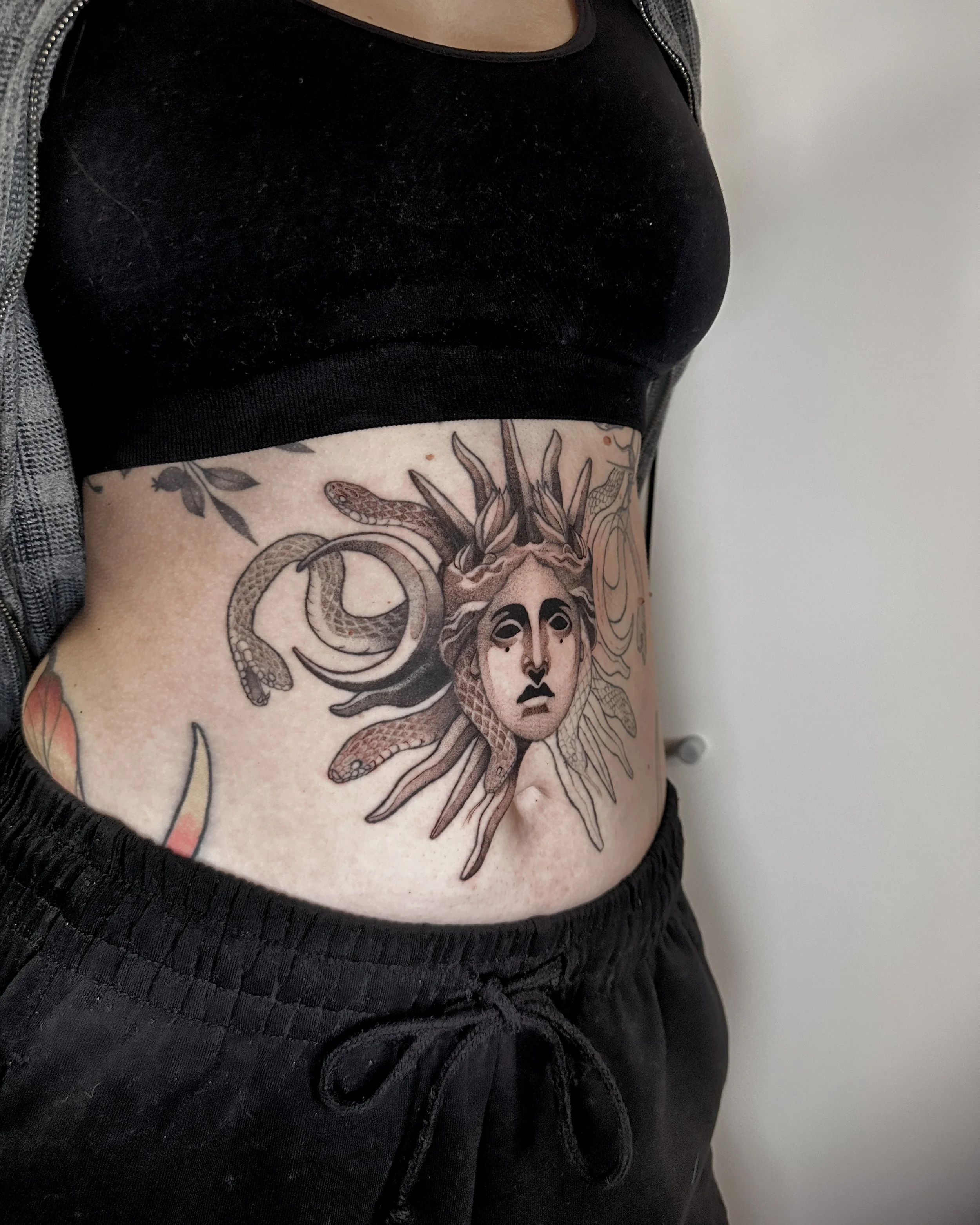 A person's side torso showing a detailed tattoo of a girl with flowing hair, snakes intertwined around her, and leaves, while wearing a black sleeveless top and a gray outer garment. its a medusa tattoo