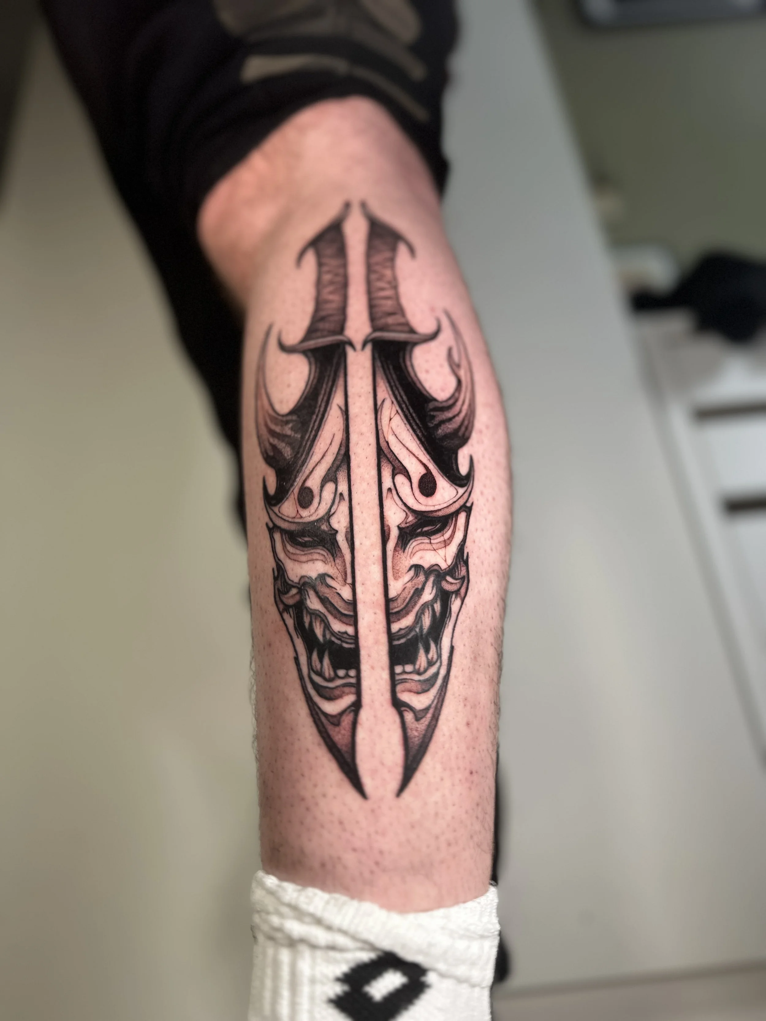 A tattoo of a symmetrical dagger with ornate details on a person's leg. the dagger is mixed with the face of a traditional japanese Hannya demon