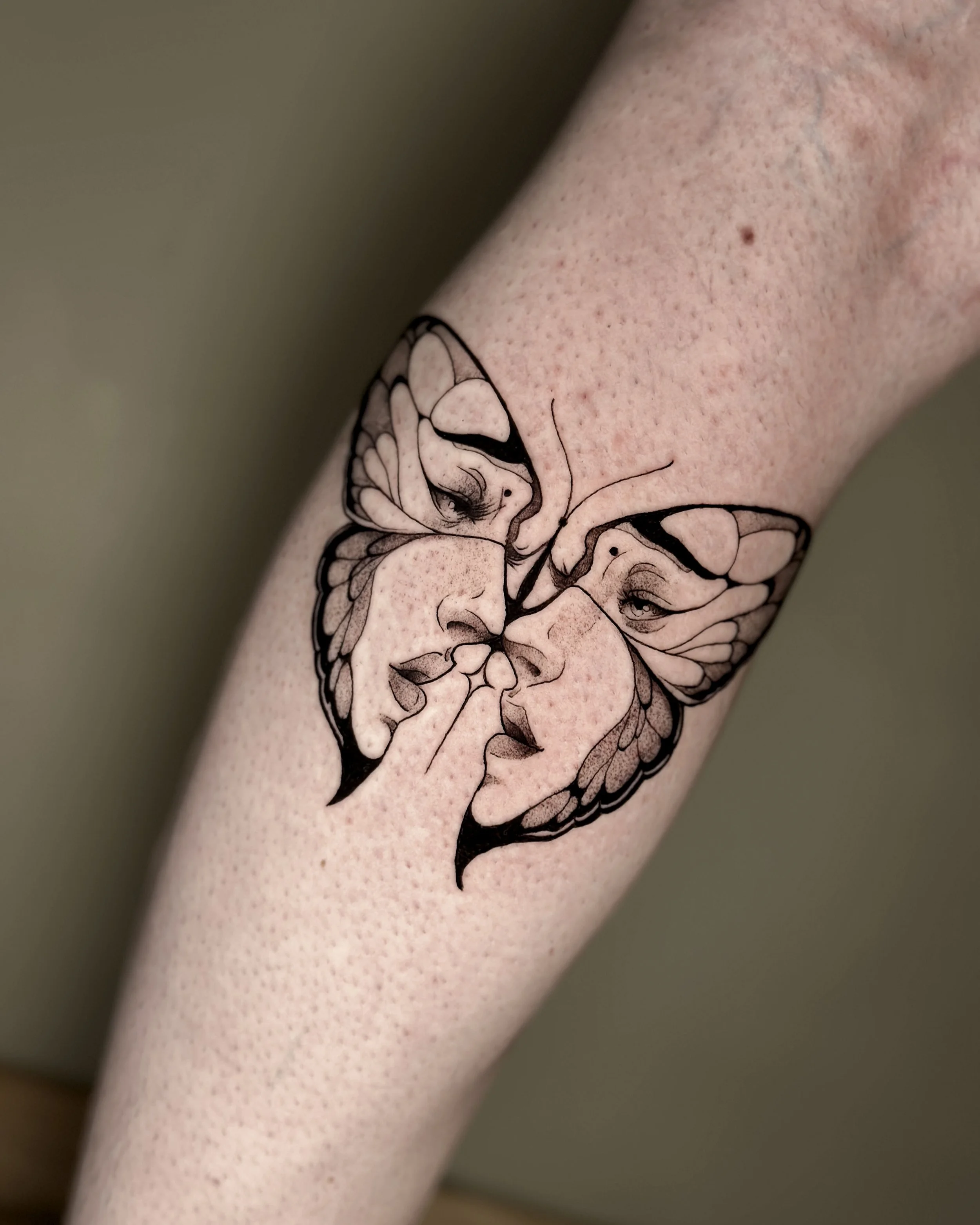 Close-up of a person's arm with a tattoo of a butter fly with 2 faces in it, with detailed line art of the butterfly's wings and body.