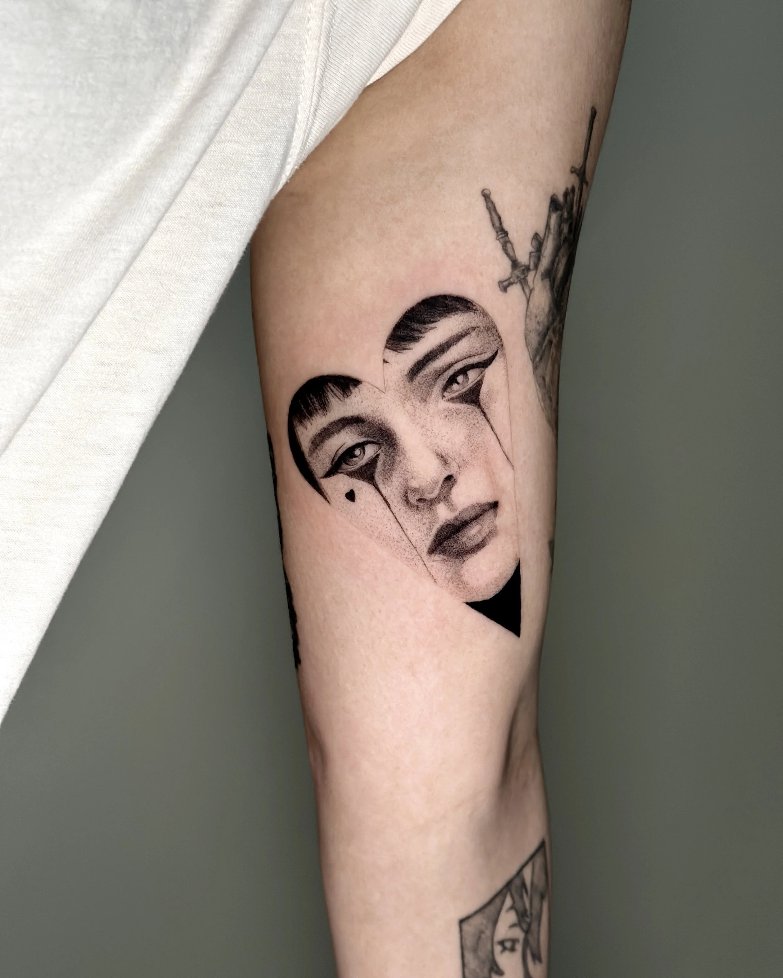 Close-up of a person's upper arm with tattoos, including a portrait, a skull, and other designs, against a gray heart shaped background.