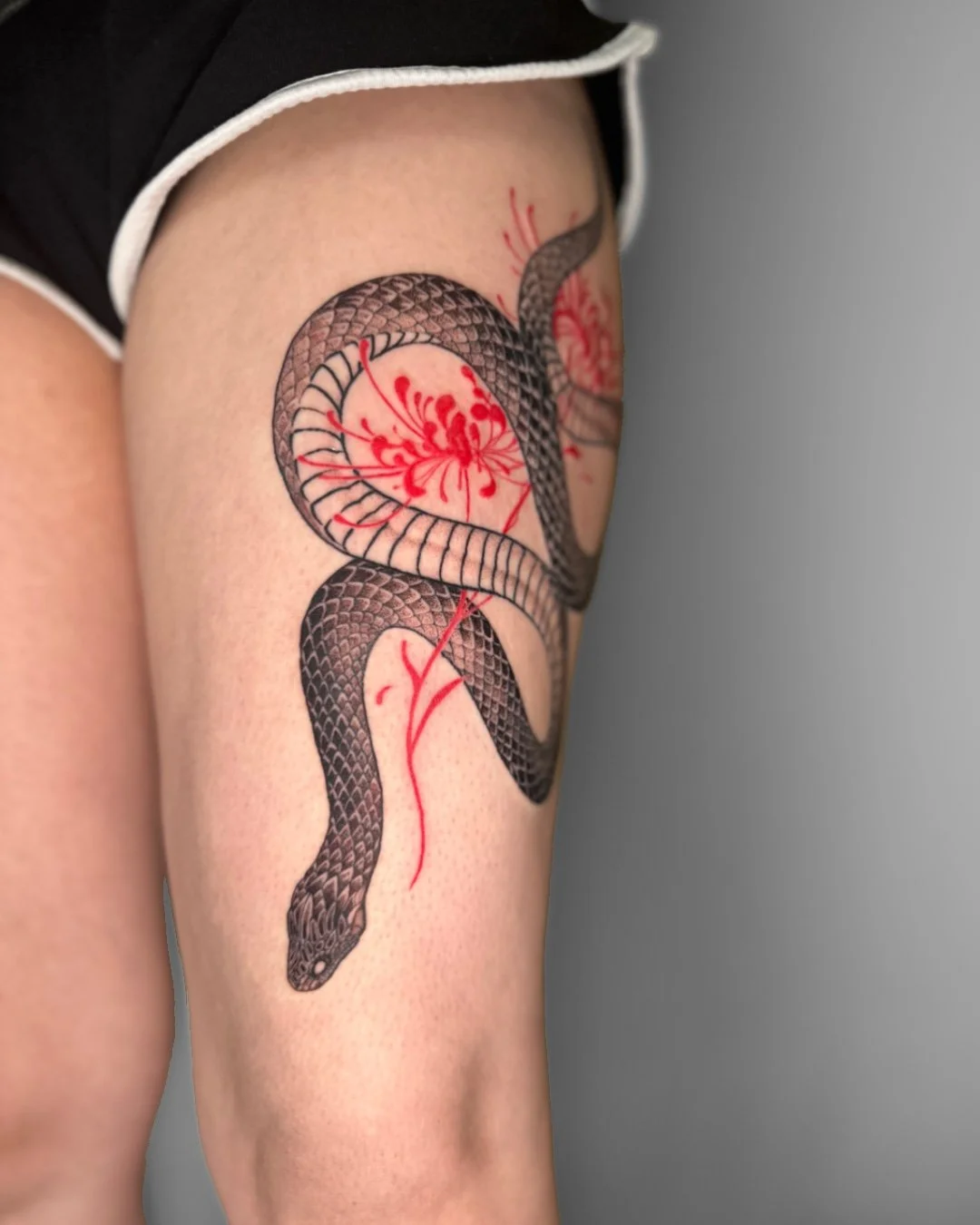 Tattoo of a black snake with detailed scales intertwined with a red flower on a person's upper thigh.