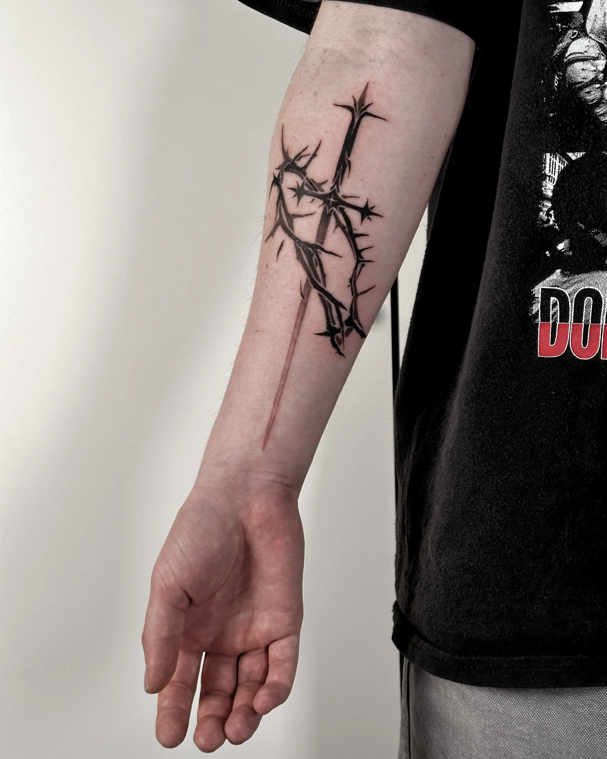 A person's left hand and arm with a black tattoo of a sword surrounded by a crown of thornes in a abstract, stylized design. The person is wearing a black shirt with a graphic print and light-colored pants, standing against a plain white background.