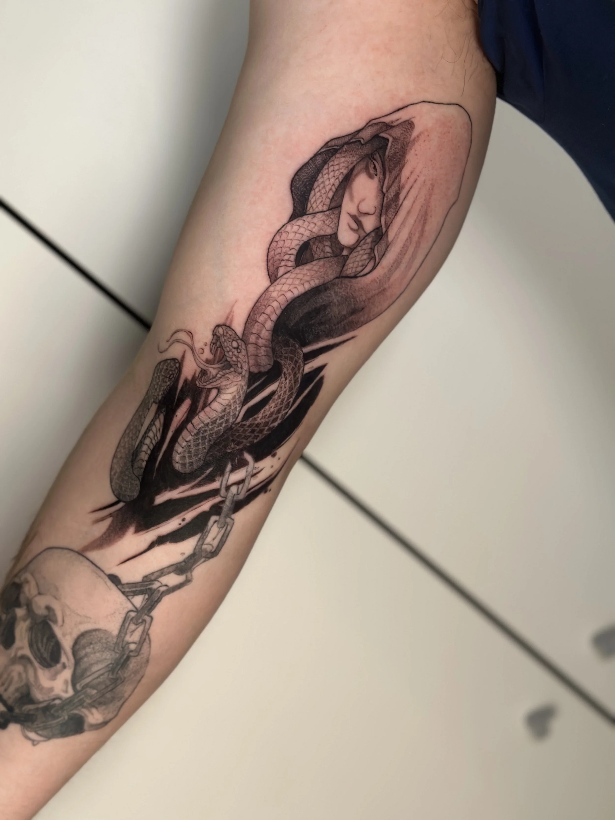 Tattoo of a woman's face with closed eyes, surrounded by snakes and chains on a person's arm. its a Medusa tattoo.
