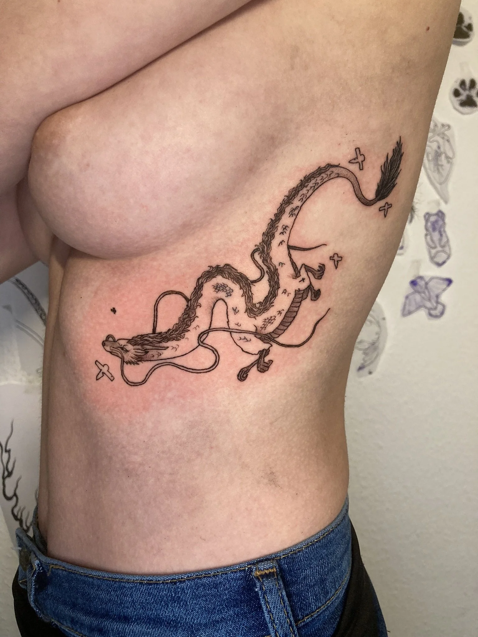 Close-up of a person's side torso showing a detailed black ink tattoo of a dragon with stars surrounding it. The dragon is Haku from Studio Ghibli's Spirited Away.