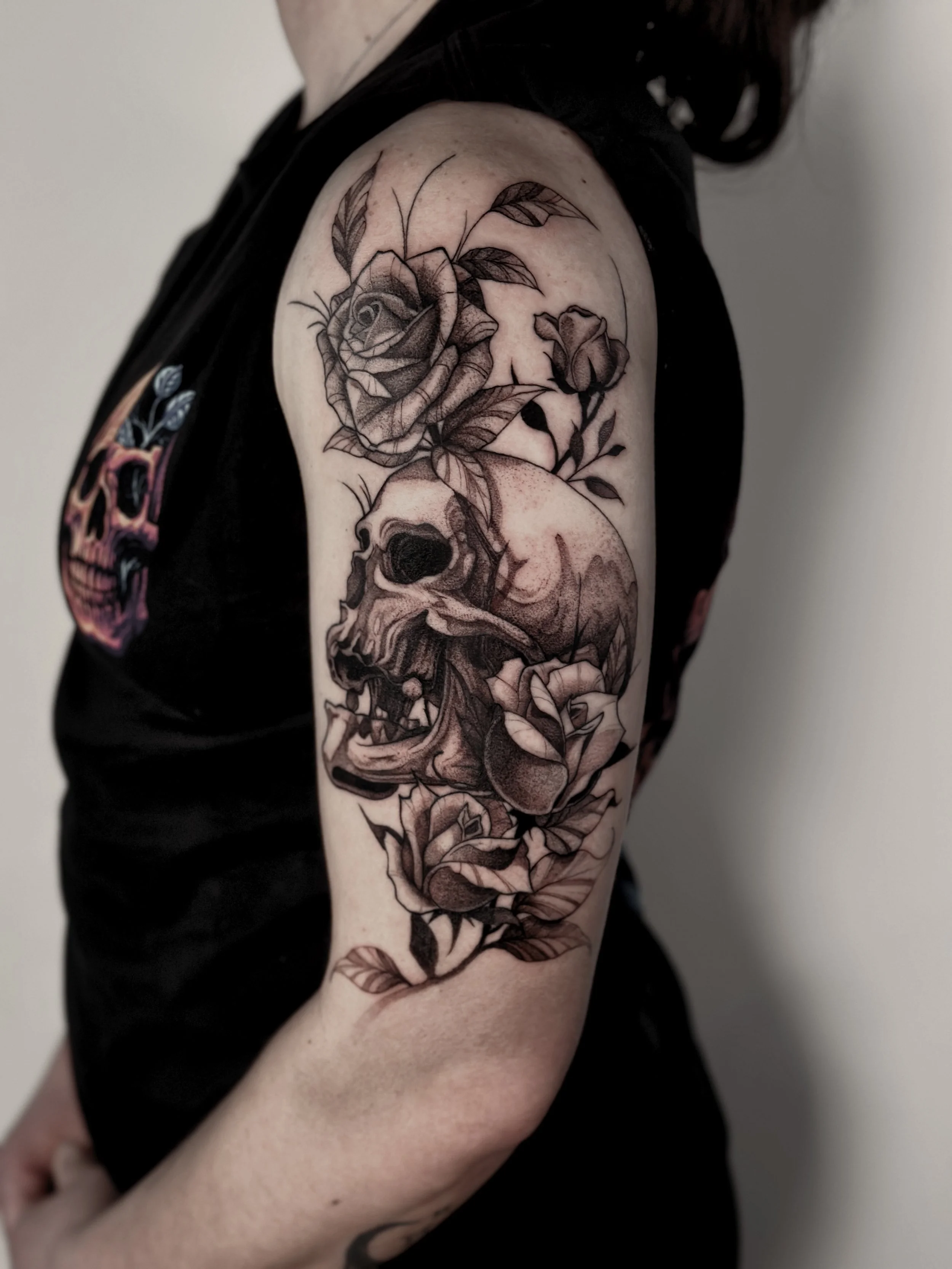 Black and gray tattoo on upper arm features a detailed rose with leaves, a skull.