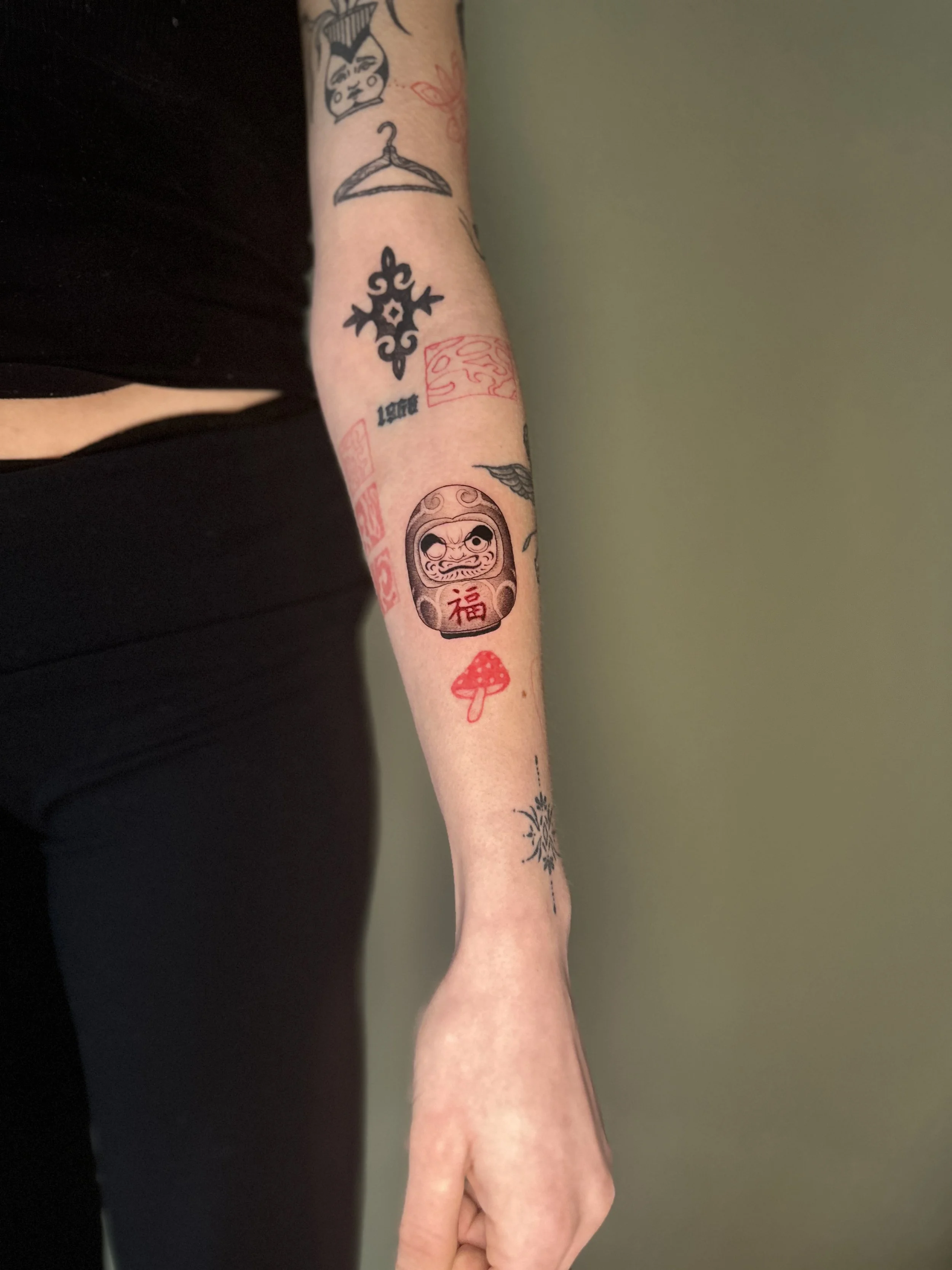 A person's arm with several tattoos, including a Daruma doll with red Japanese characters, a small mushroom, a floral pattern, and a winged design, with the person's hand relaxed and fingers gently curved.