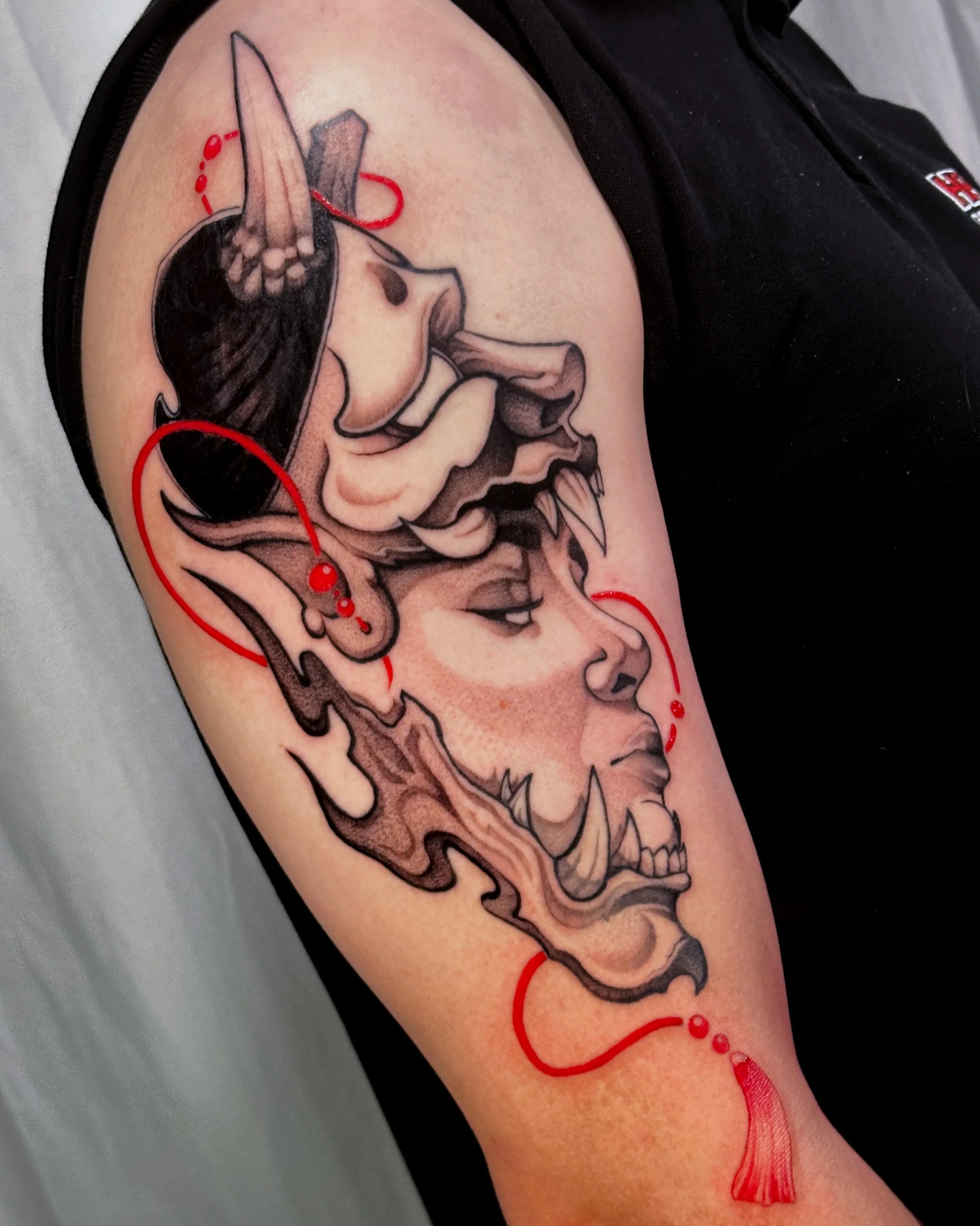 Tattoo of a woman's profile with elements of traditional japanese hannya tattoo art, including a red dotted line embellishment and a red tassel, on a person's upper arm.