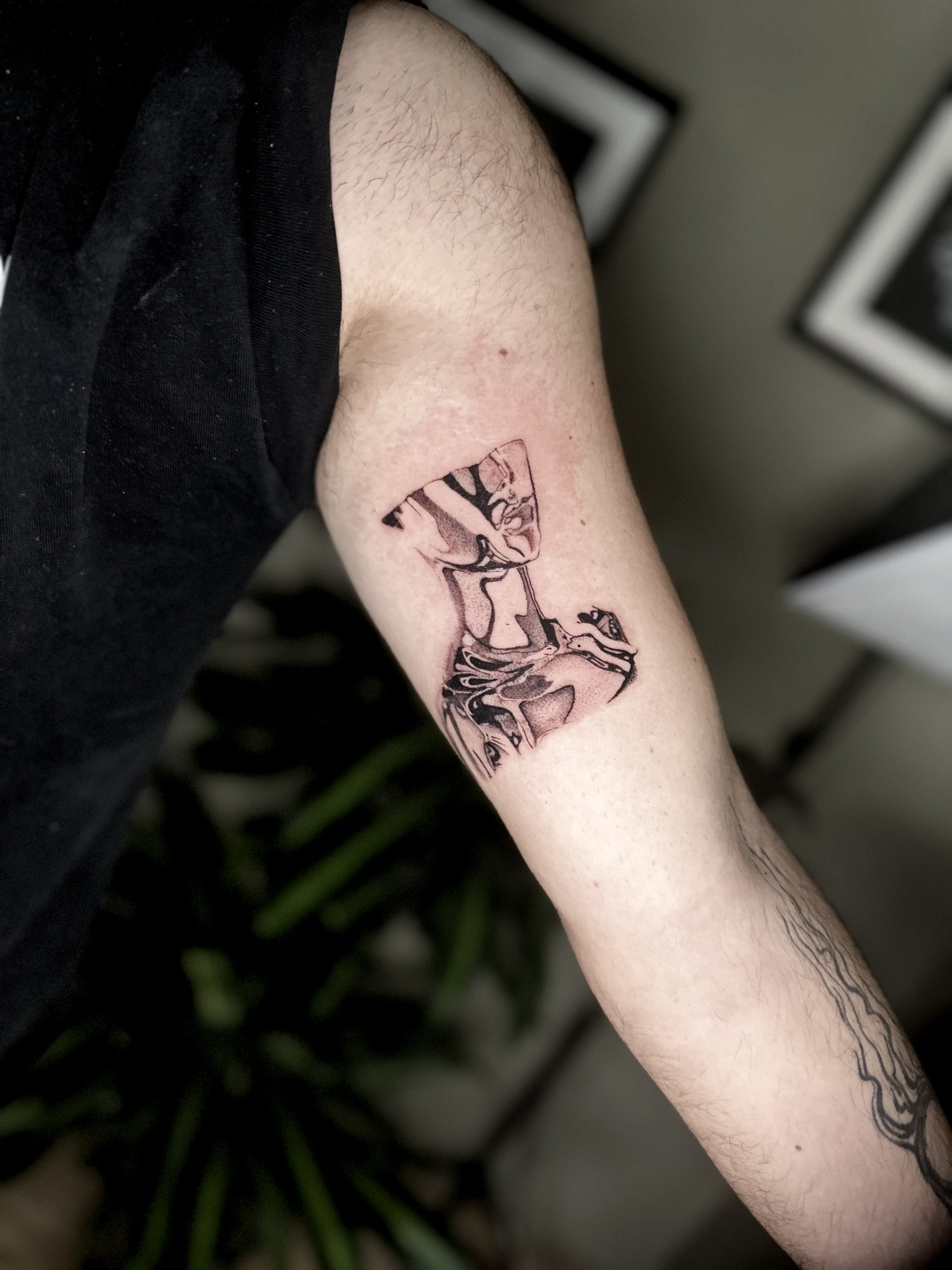 A person's forearm with a tattoo of a detailed illustration of a chromed reflective upper female body. The background includes some indoor framed pictures and a green plant.