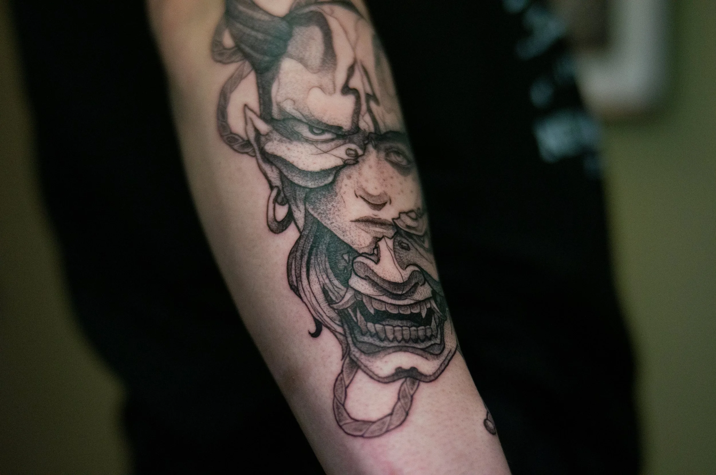 A tattoo of a woman's face with curly hair, earrings, and a confident expression on her arm mixed with a japanese hannya mask that is broken.
