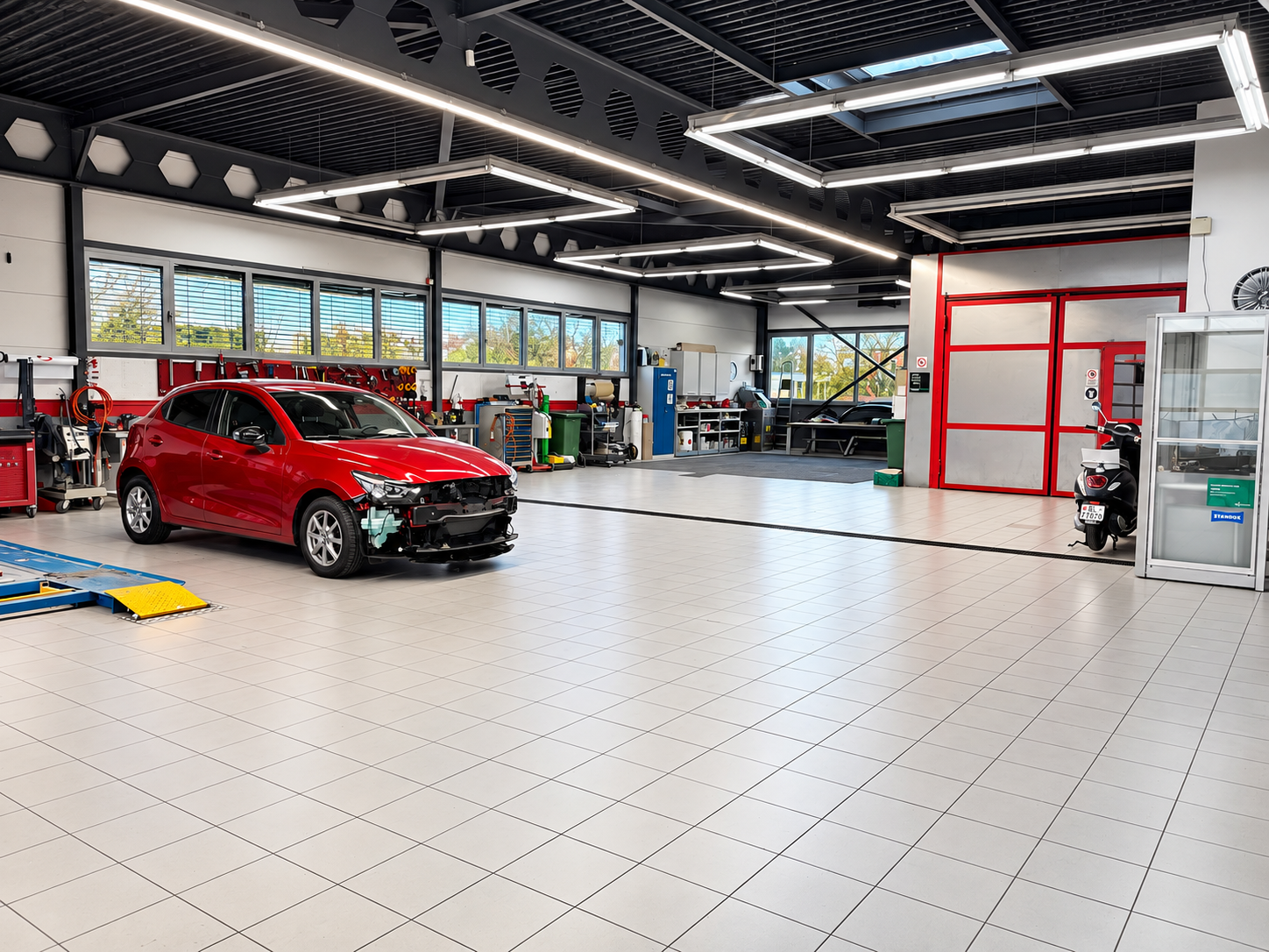 Innenraum einer Autowerkstatt mit einem roten Auto, das repariert wird, und verschiedenen Werkzeuge und Ausrüstung.
