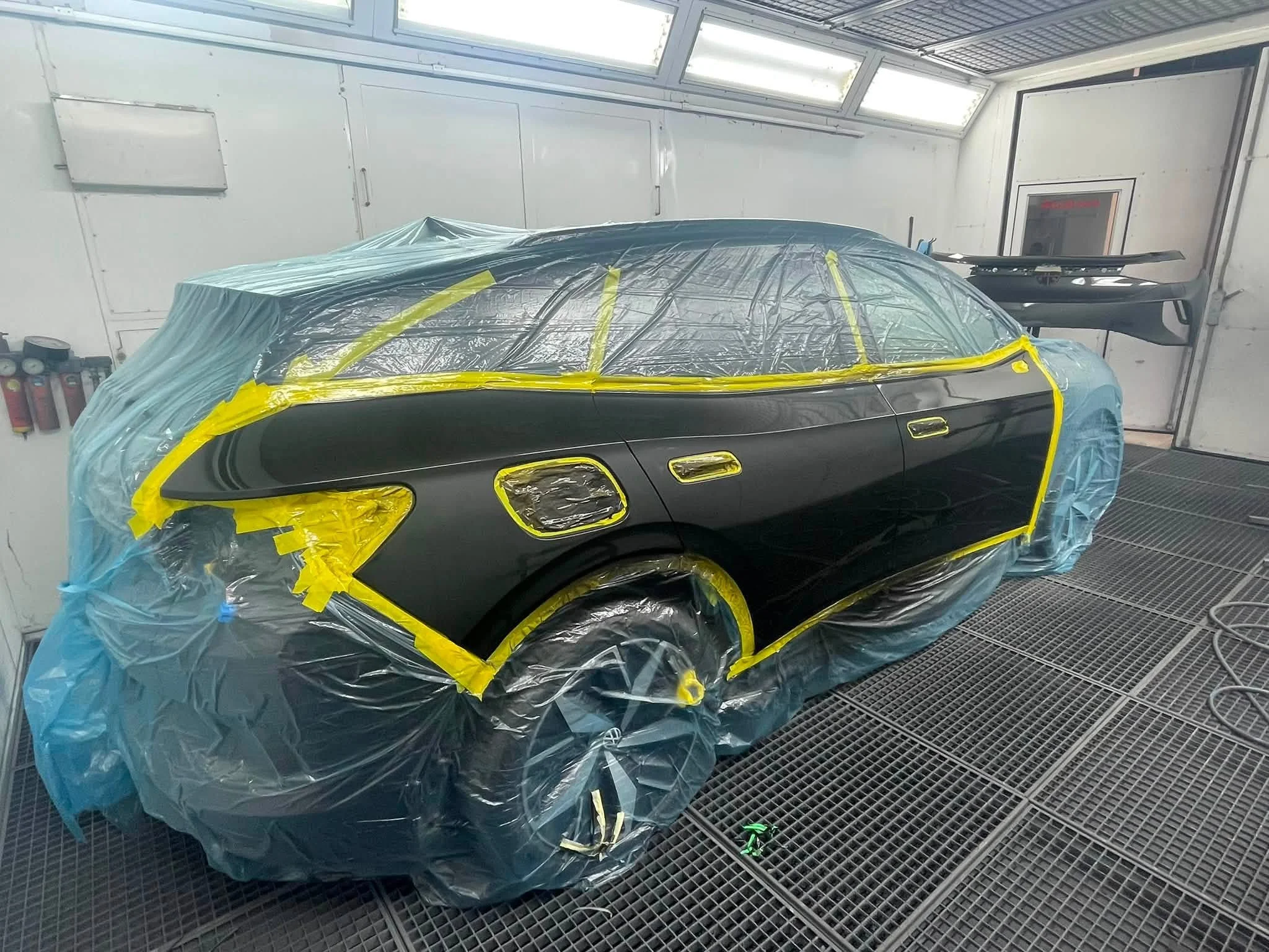Ein schwarzes Auto wird in einer Werkstatt lackiert, eingewickelt in blaue und gelbe Abdeckfolien. Das Fahrzeug befindet sich in einem professionellen Lackierraum mit weißen Wänden und einer Gitterboden.