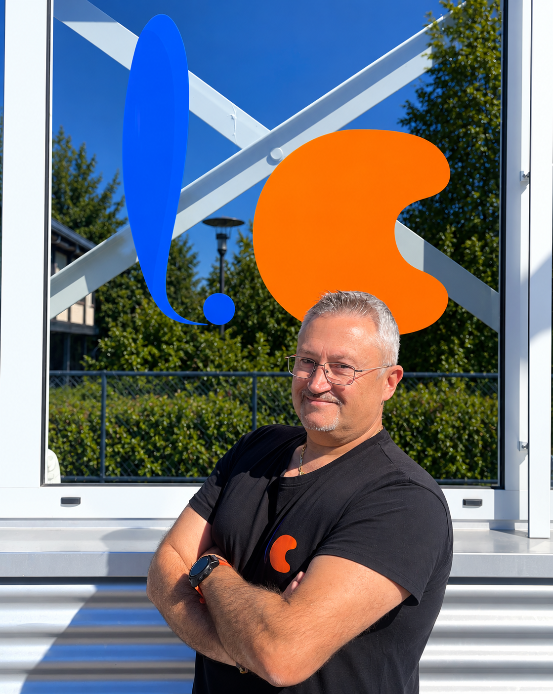 Ein Mann steht vor einer großen Glaswand mit farbigen abstrakten Symbolen, trägt ein schwarzes T-Shirt mit einem kleinen orangefarbenen Logo, hat kurz graues Haar, trägt Brille und eine Armbanduhr.