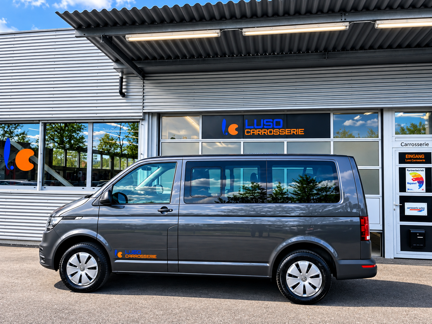 Ein grauer Van mit Luso Carrosserie Logo vor einer Werkstatthalle mit gleichen Logos, reflektierende Fenster, blauer Himmel mit Wolken, grüne Bäume, Asphaltboden.