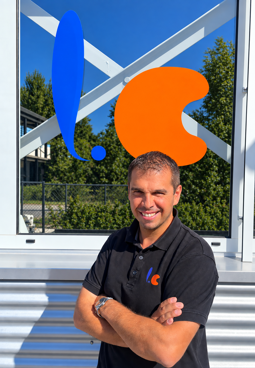 Ein Mann in einem schwarzen Polo-Shirt mit einem Logo steht vor einem großen Glasschirm mit einem bunten Logo, im Hintergrund Bäume und ein Zaun bei sonnigem Wetter.