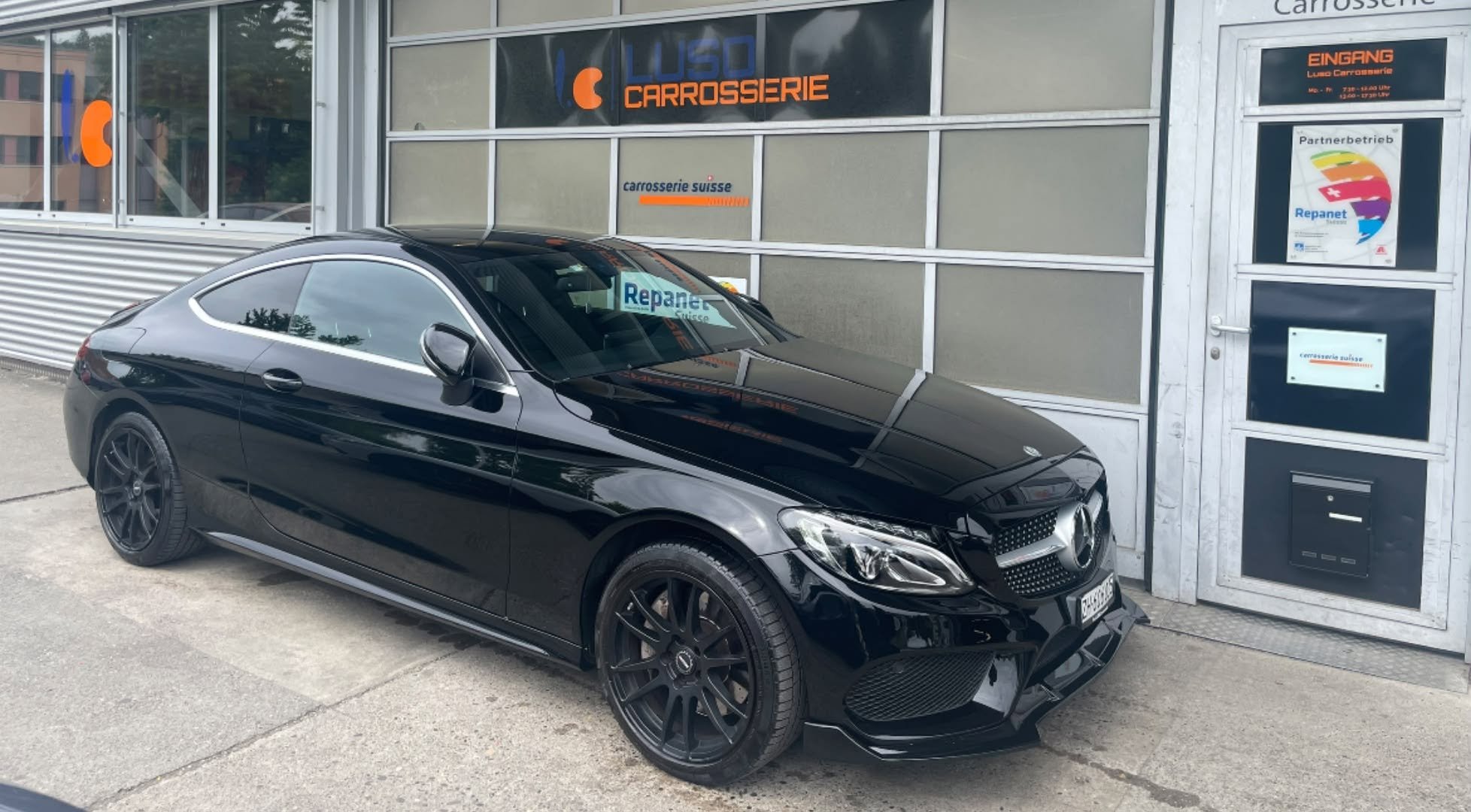 Schwarzer Mercedes-Benz in der Nähe eines Autohauses mit Schaufenstern, Fenstern und Türen. Die Fenster tragen das Logo und den Namen des Autohauses. Das Auto ist modern und sportlich gestaltet, mit schwarzen Rädern und einem schwarzen Lack.