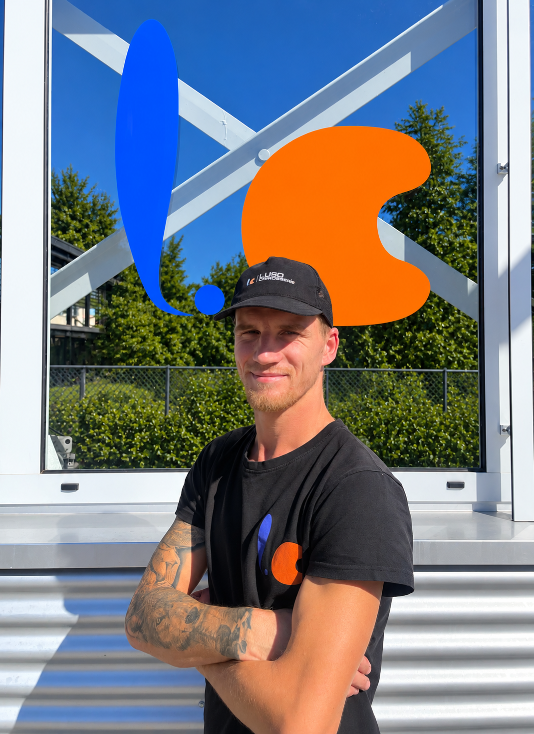 Ein junger Mann mit Tattoos trägt ein schwarzes T-Shirt und eine schwarze Kappe mit dem Logo 'Luso' vor einem Glas mit einer blauen und orangenen Grafik.