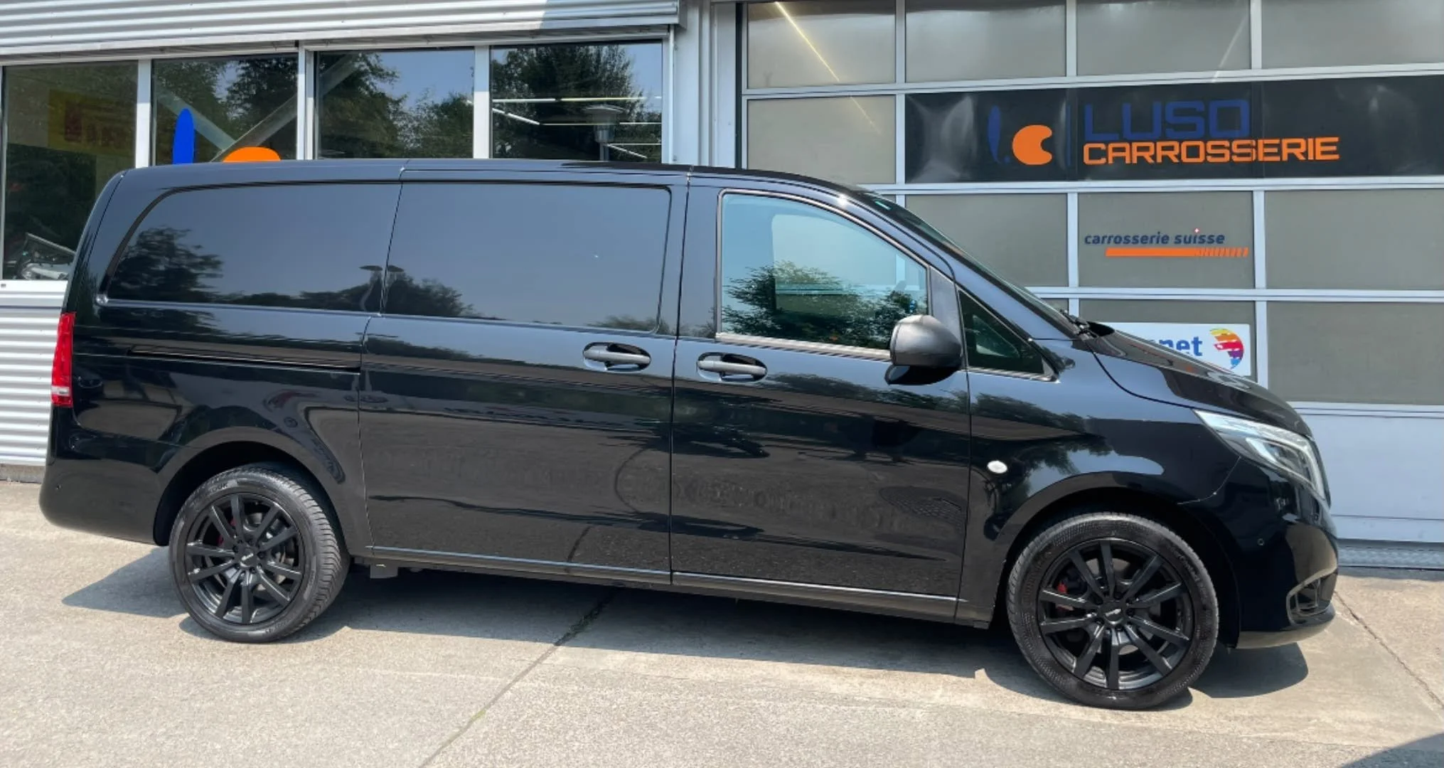Schwarzer Van parkt vor einem Geschäft mit Glasfenstern, das Schilder mit den Worten 'Luso Carrosserie' und 'Carrosserie Suisse' hat.
