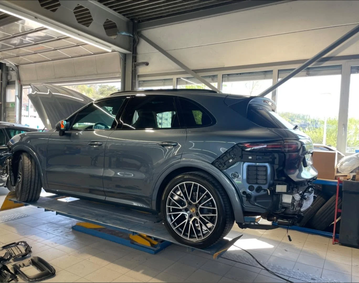 Ein schwarzer Porsche SUV befindet sich in einer Werkstatt, teilweise zerlegt mit offenem hinteren rechten Rad und abgebautem frontseitigem Bauteil.