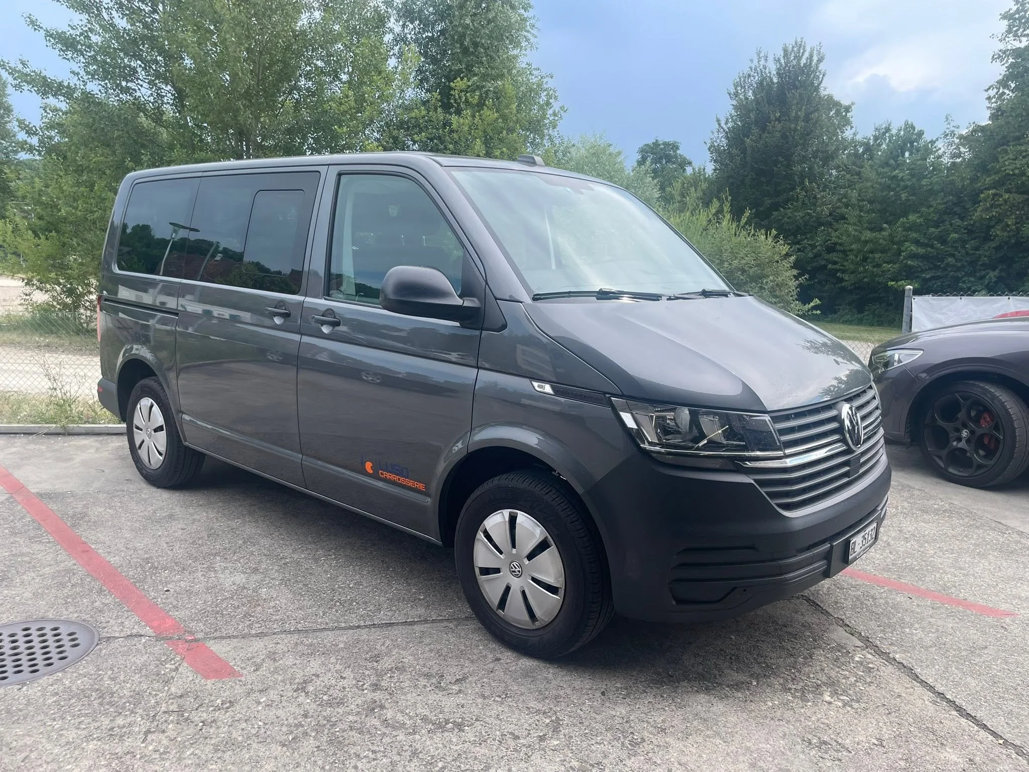 Ein grauer Transporter der Marke Volkswagen steht in einer Parkbucht auf einem Parkplatz, im Hintergrund sind Bäume und ein blauer Himmel sichtbar.