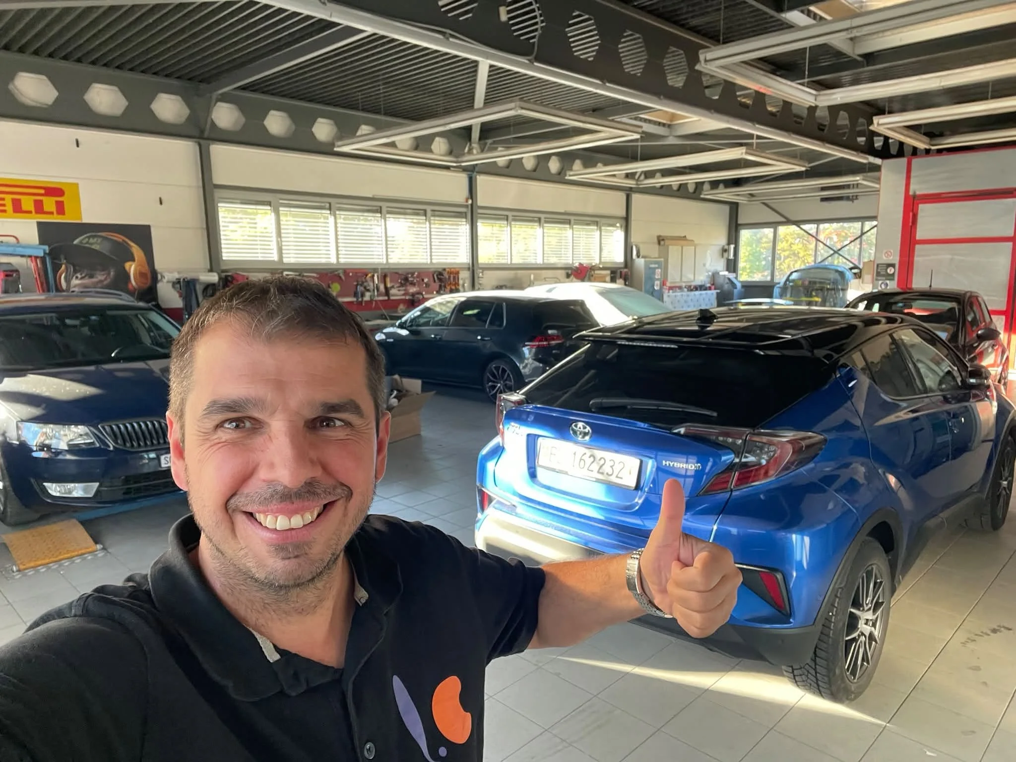 Ein Mann lächelt in einer Garage mit mehreren geparkten Autos und hebt den Daumen. Im Hintergrund sind Werkzeuge und eine Werkbank sichtbar.
