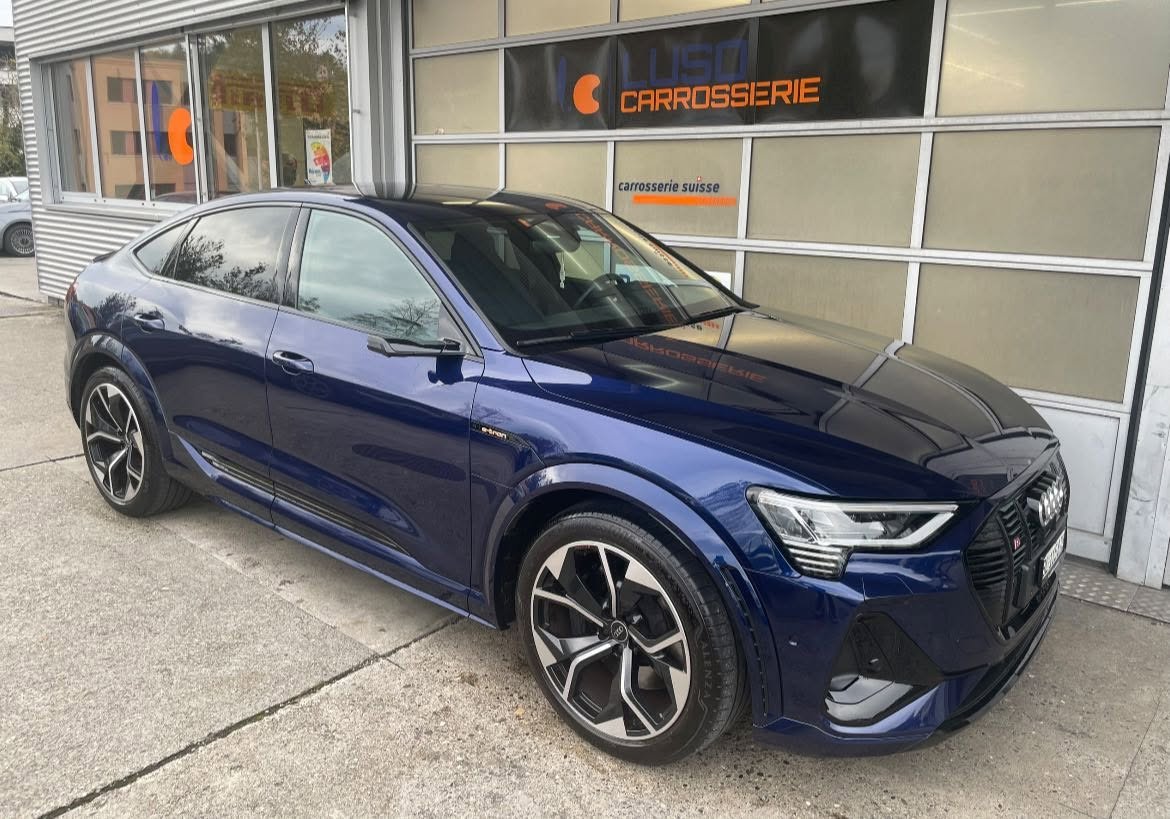 Ein blauer moderner Audi E-Tron Quattro Standauto vor einem Carrosserie-Geschäft mit der Beschriftung −LUSO Carrosserie− und −carrosserie suisse− an einem grauen Tag, aufgenommen von außen.