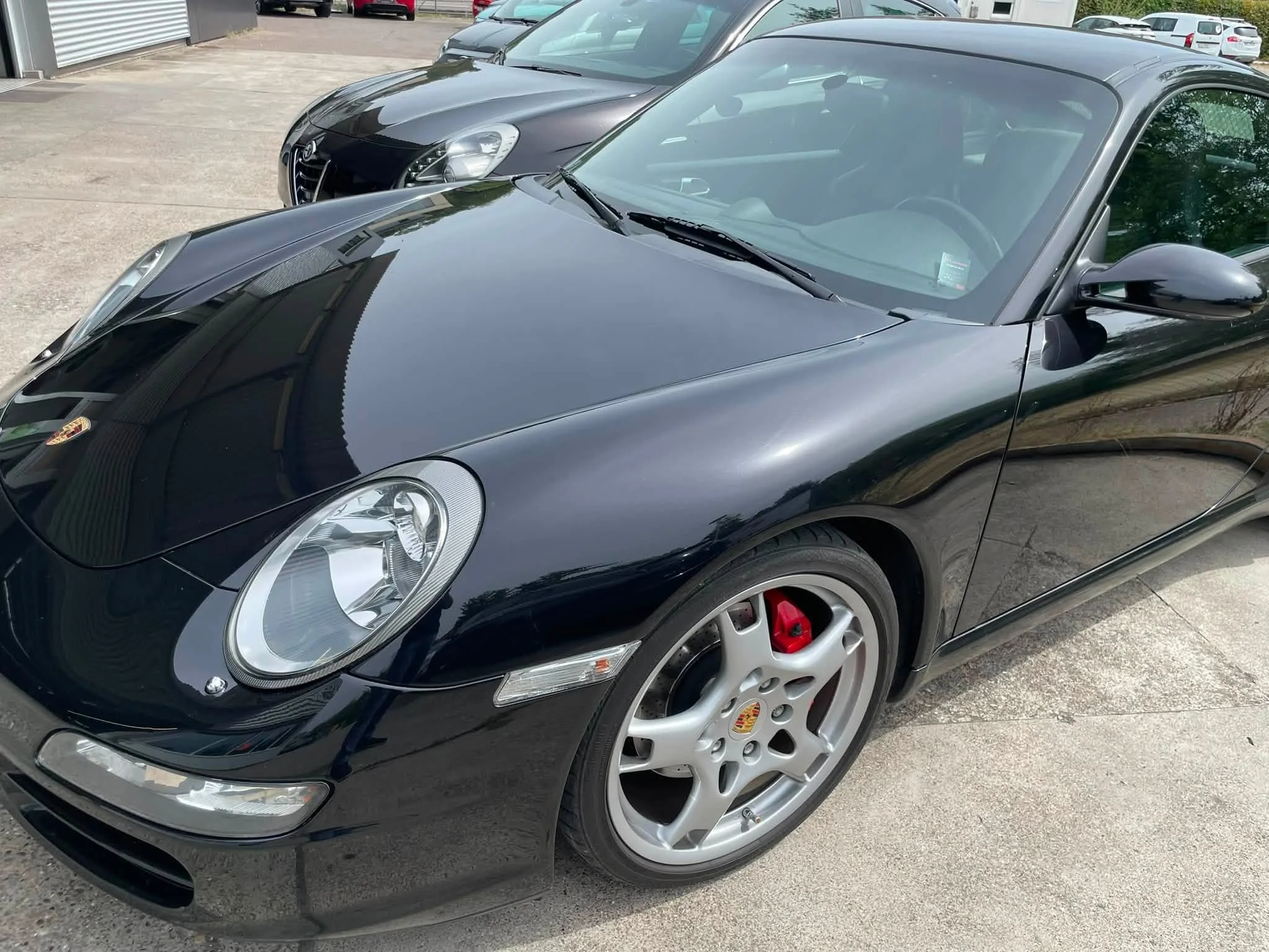 Zwei schwarze Autos, eines Porsche und eines Alfa Romeo, parken auf einem Parkplatz bei sonnigem Wetter.
