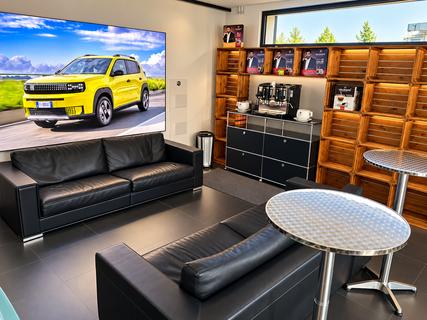 Wartebereich mit schwarzen Ledersofas, runden Metalltischen, einem großen Fernseher, einem Kaffeeautomaten und leeren Holzregalen, Fenster mit Blick auf Bäume im Hintergrund.