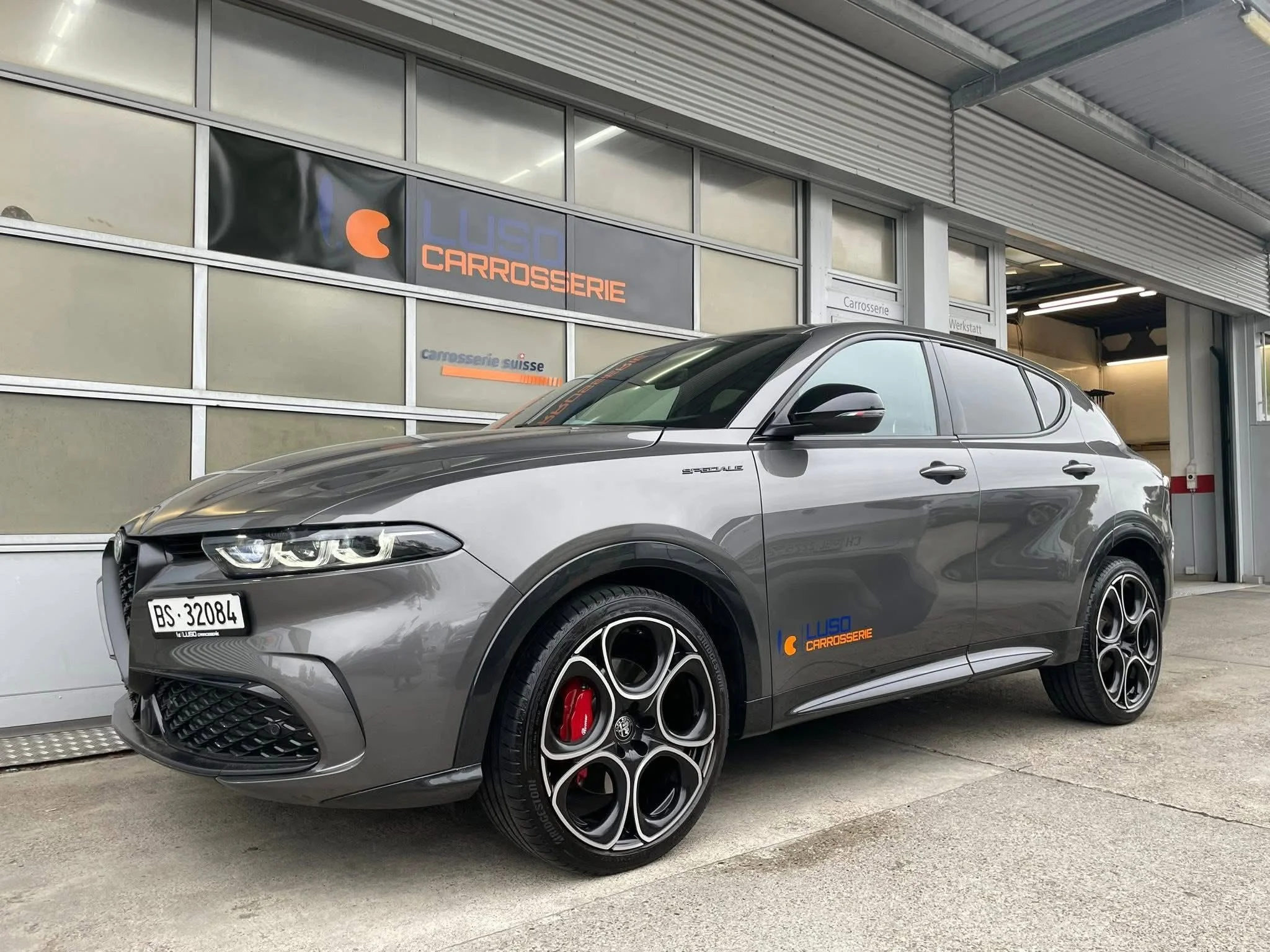 Ein grauer Sportwagen steht vor einer Werkstatt mit Schildern, die 'Carrosserie' und 'Werkstatt' anzeigen, im Vordergrund. Das Fahrzeug hat eine sportliche Optik mit schwarzen Felgen und roten Bremsen, und trägt ein Kennzeichen mit der Nummer BS 3208