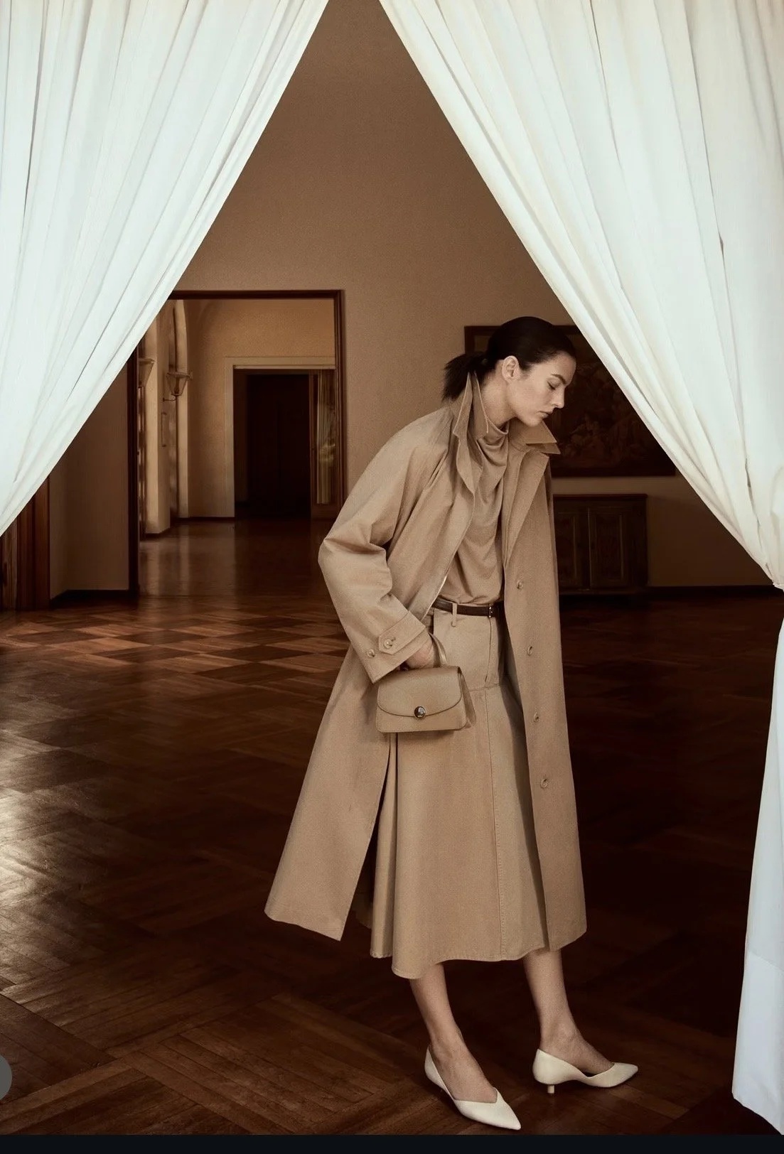 A woman in beige clothing stands under white curtains in a room with wooden floors and beige walls, looking down with her hands in her coat pockets, holding a small beige handbag.