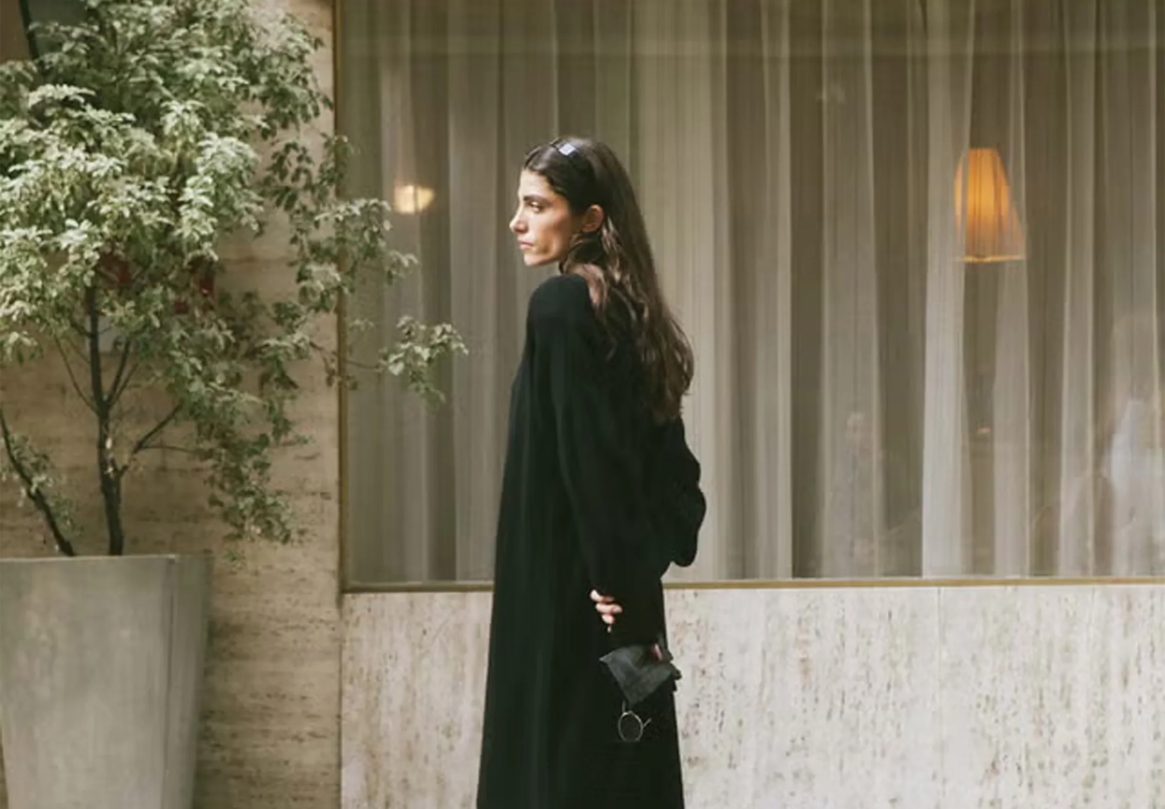A woman with long, wavy dark hair, wearing a black outfit, standing indoors next to a tree with white blossoms, with beige curtains and a wall-mounted lamp in the background.