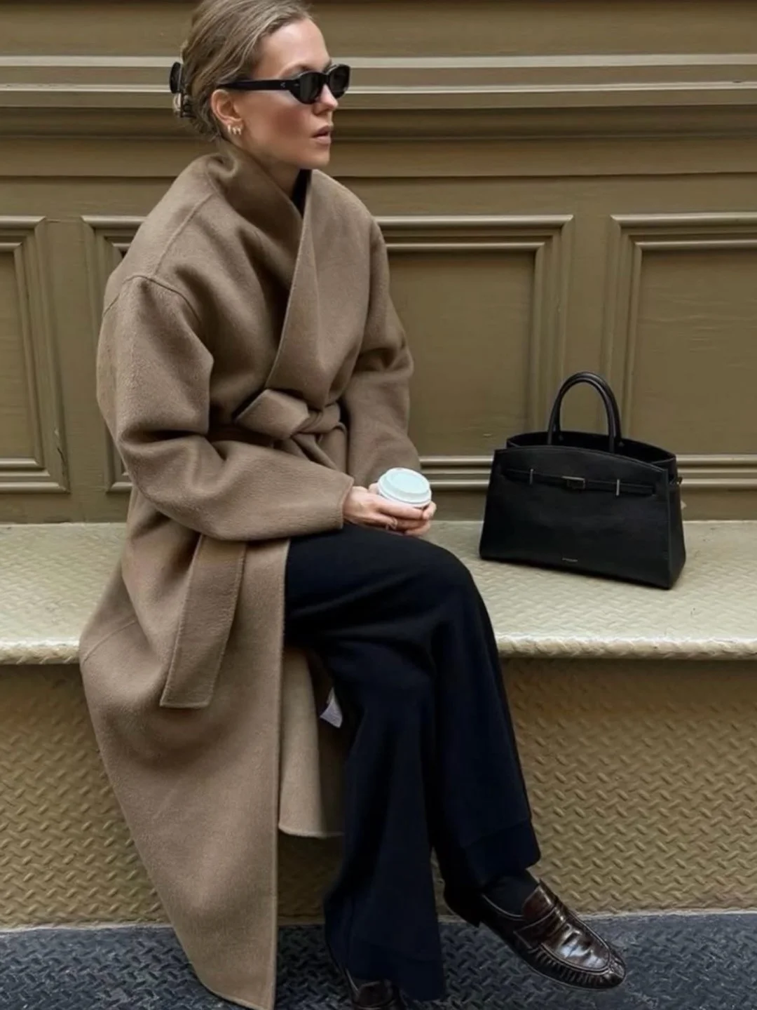A woman sitting on a bench in a waiting area, wearing sunglasses, a beige coat, black pants, and loafers, holding a coffee cup, with a black handbag placed next to her.